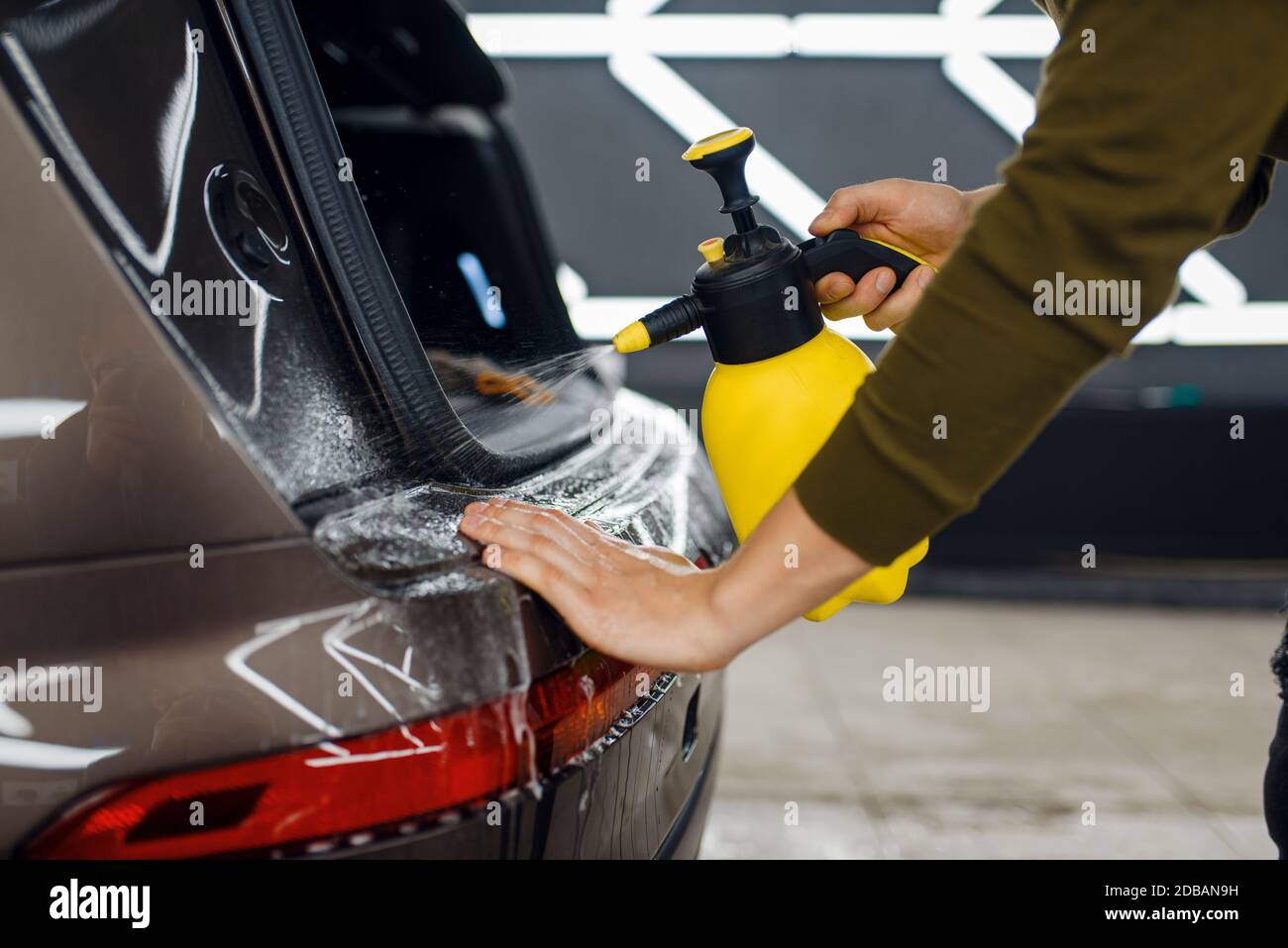 Il lavoratore maschio bagna la superficie del paraurti posteriore dell'auto con spray prima di applicare la pellicola di protezione. Installazione del rivestimento che protegge la vernice di automobi Foto Stock