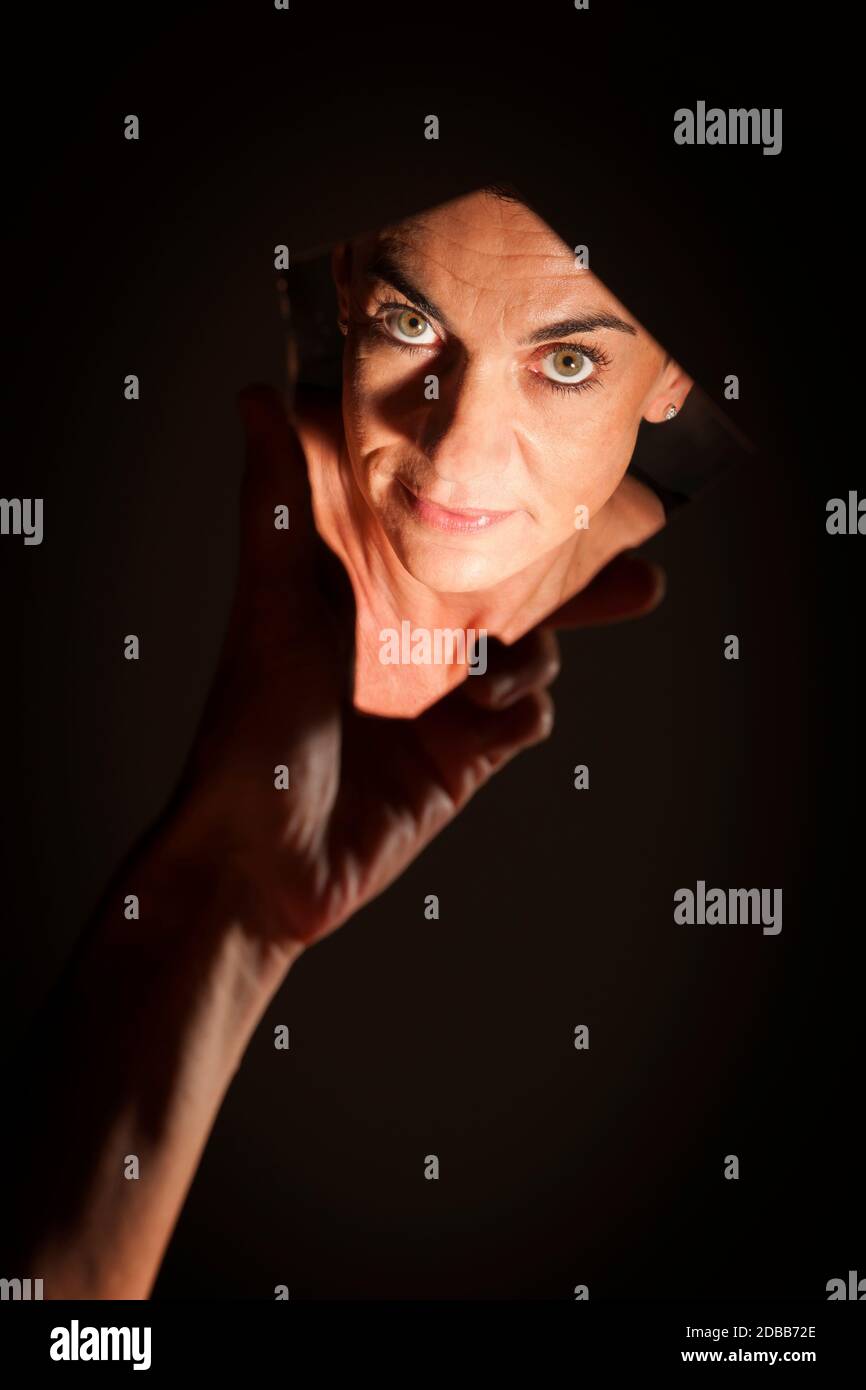 Ritratto facciale di una donna dai capelli neri di mezza età che guarda nella macchina fotografica in uno specchio a mano che brulica su uno sfondo scuro. Foto Stock
