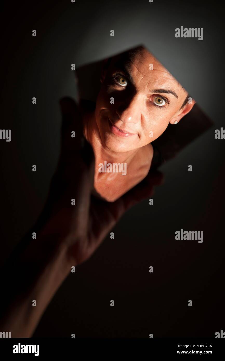 Ritratto facciale di una donna di mezza età dai capelli neri che guarda nella fotocamera in uno specchio tenuto a mano che si stira nella luce laterale su uno sfondo scuro. Foto Stock