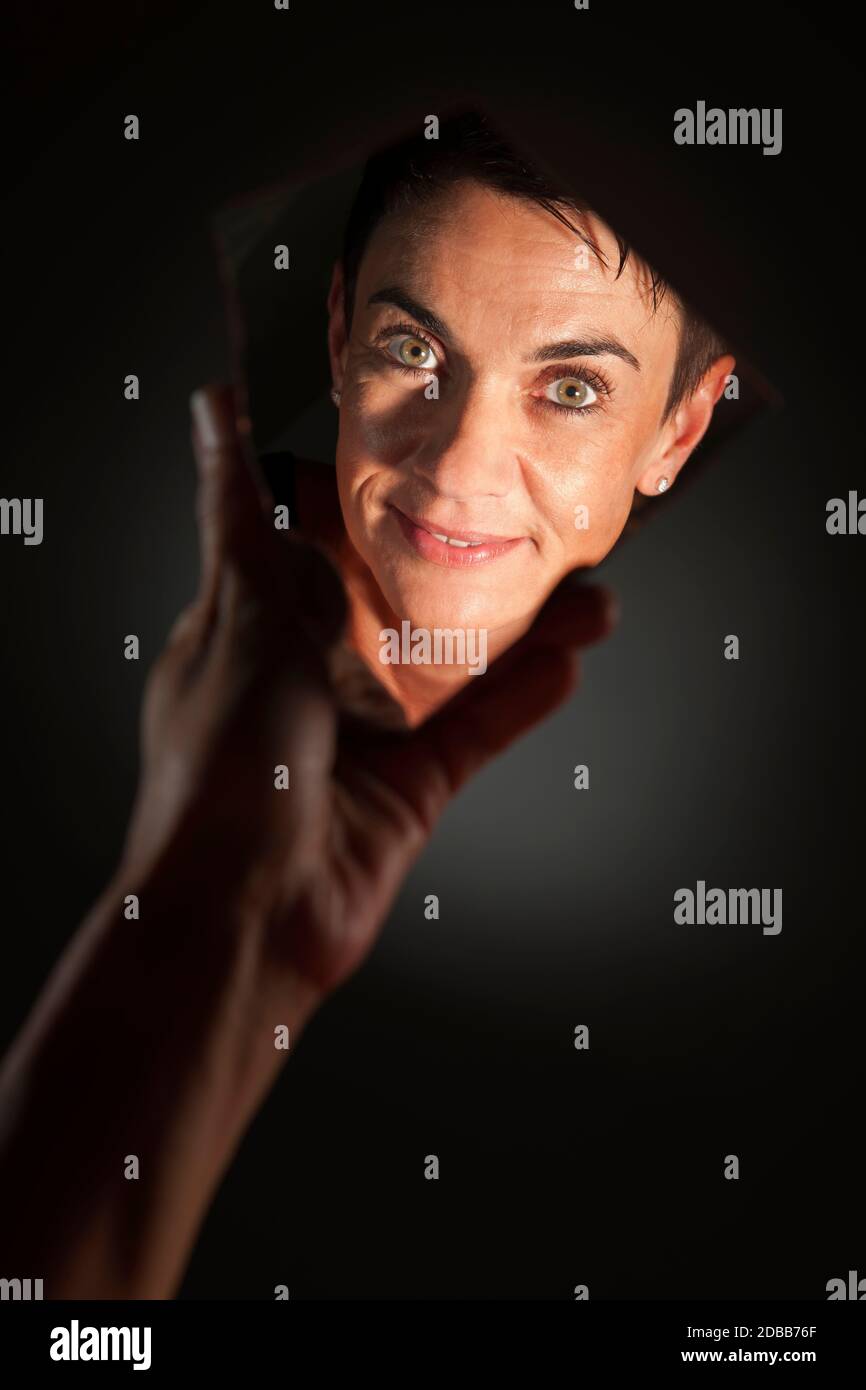 Ritratto facciale di una donna dai capelli neri di mezza età che guarda nella macchina fotografica in uno specchio a mano che brulica su uno sfondo scuro. Foto Stock