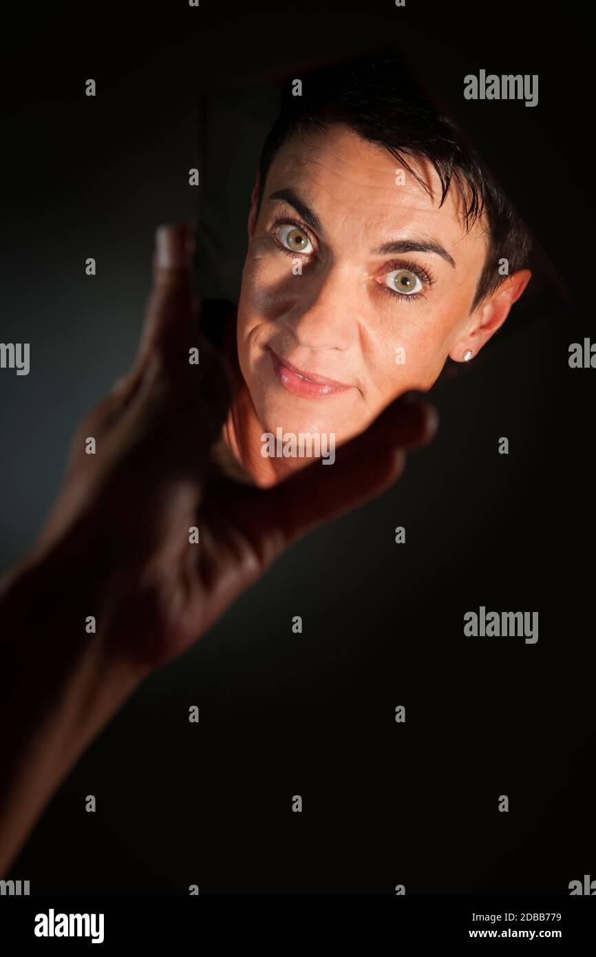 Ritratto facciale di una donna dai capelli neri di mezza età che guarda nella macchina fotografica in uno specchio a mano che brulica su uno sfondo scuro. Foto Stock