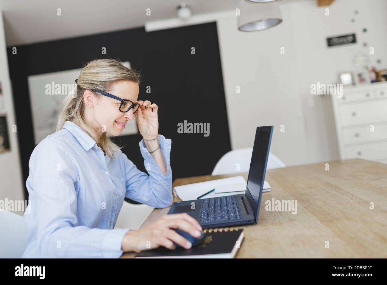 bella giovane donna con camicia blu e occhiali che fa ufficio domestico seduto al tavolo da cucina di fronte al suo laptop Foto Stock