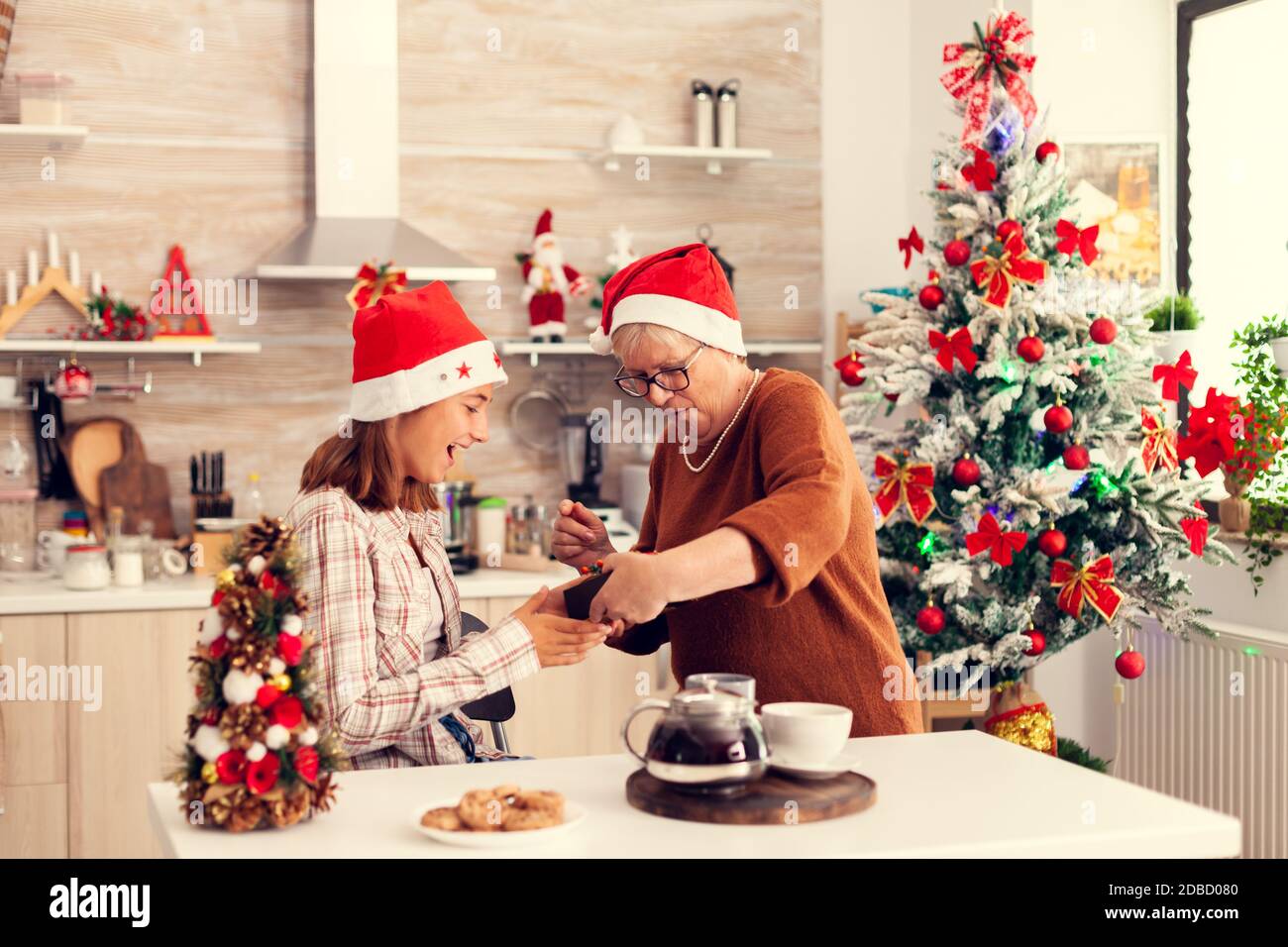 Buona nonna che dà scatola regalo della nipote durante la celebrazione di natale che indossa il cappello rosso di santa. Donna anziana che indossa il cappello di santa nipote sorprendente con la vacanza invernale presente in casa cucina con albero di Natale sullo sfondo. Foto Stock