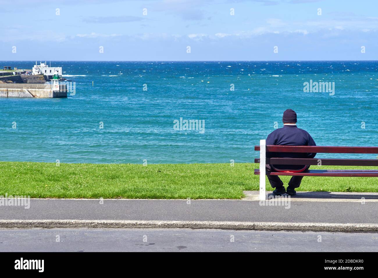 Uomo medio anziano seduto sull'estrema sinistra di una panchina guardando verso il mare in una giornata soleggiata ma fredda Foto Stock