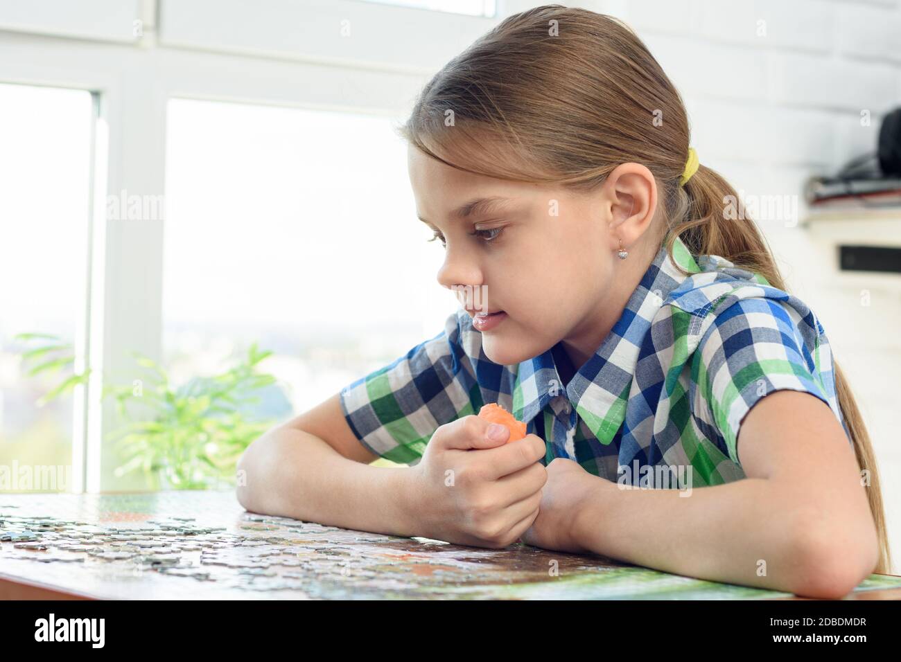 Ragazza raccoglie un puzzle mentre siede a casa al tavolo e mangia carote Foto Stock