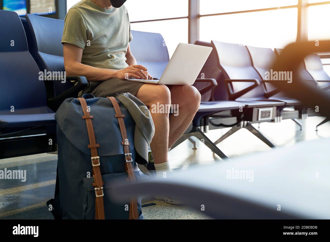 uomo asiatico viaggiatore d'aria maschile con maschera nera seduta in un'area di attesa nell'edificio del terminal dell'aeroporto utilizzando un computer portatile Foto Stock
