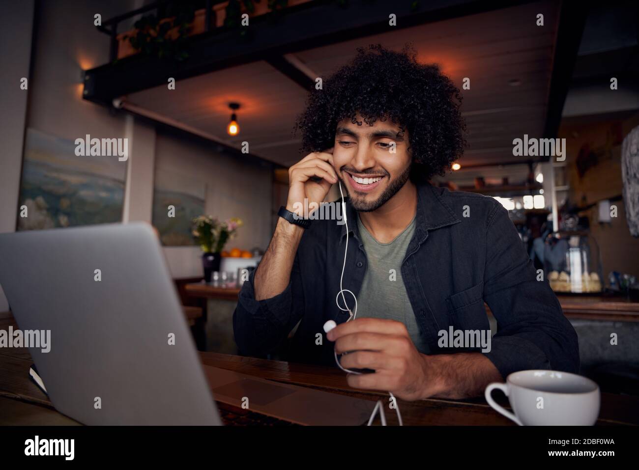 Bell'uomo giovane con capelli ricci che guarda i video sul computer portatile con auricolari in caffetteria Foto Stock