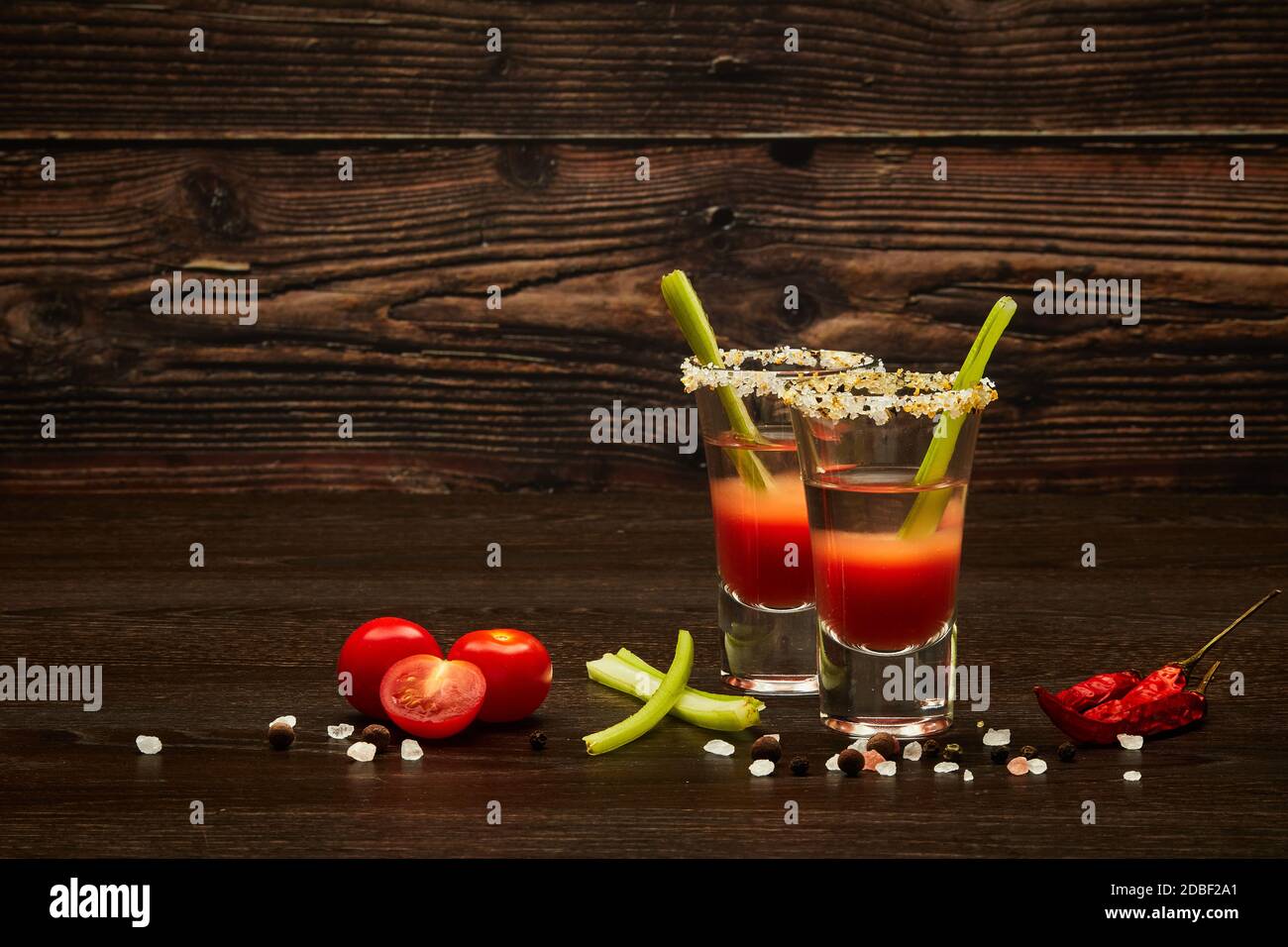 Due bicchieri da shot con cocktail Bloody Mary, sedano, spezie e pomodori maturi su sfondo di legno scuro con spazio per la copia Foto Stock