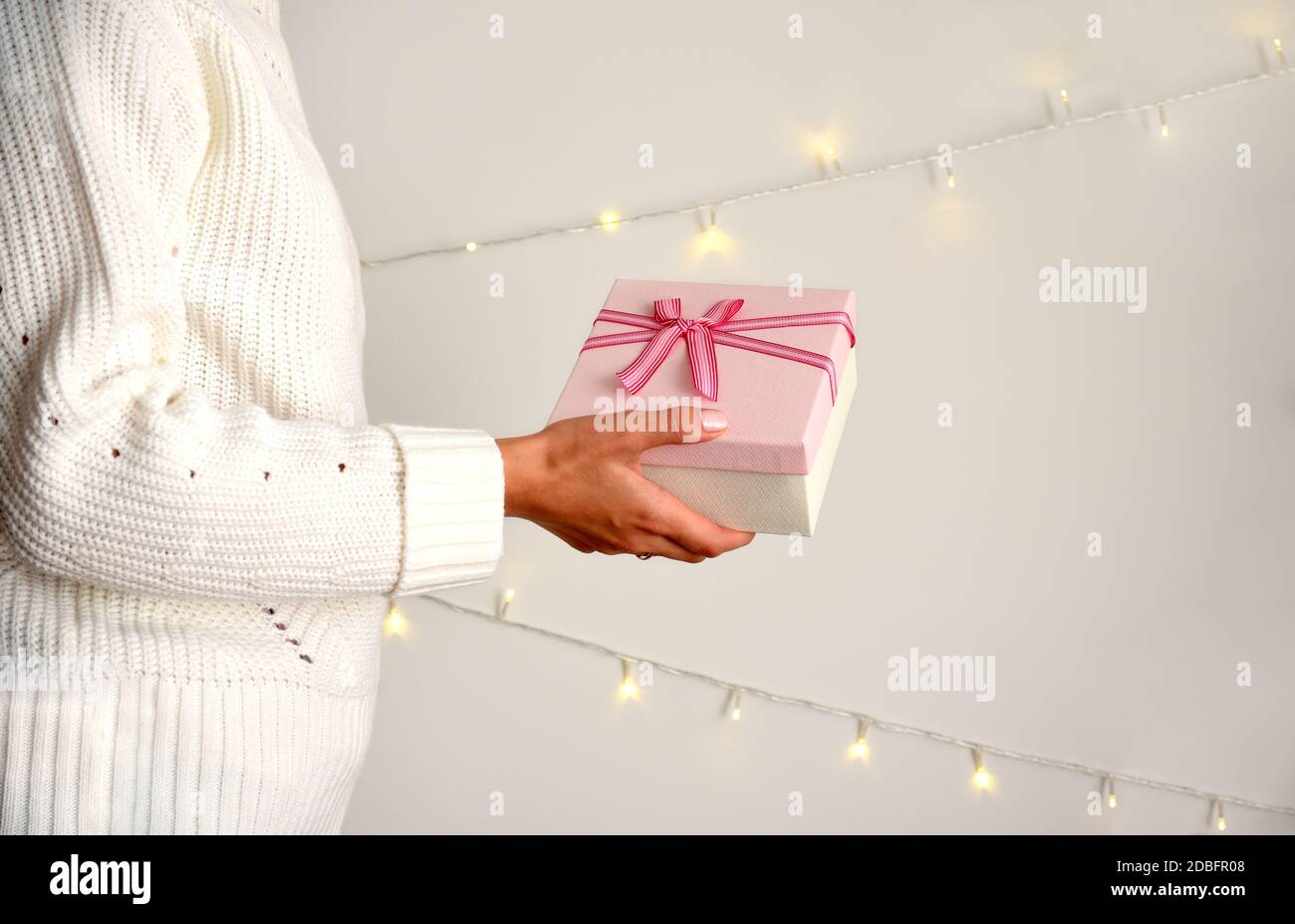 Confezione regalo rosa con prua in mano femminile in pullover bianco su sfondo chiaro. Ghirlande di Natale sul muro Foto Stock