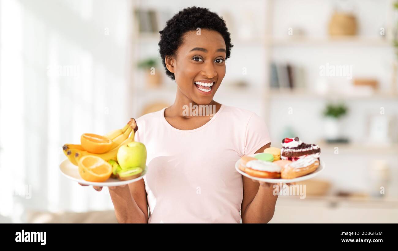 Donna nera felice che tiene piatti con frutta e dessert a casa, facendo la scelta tra cibi sani e malsani Foto Stock