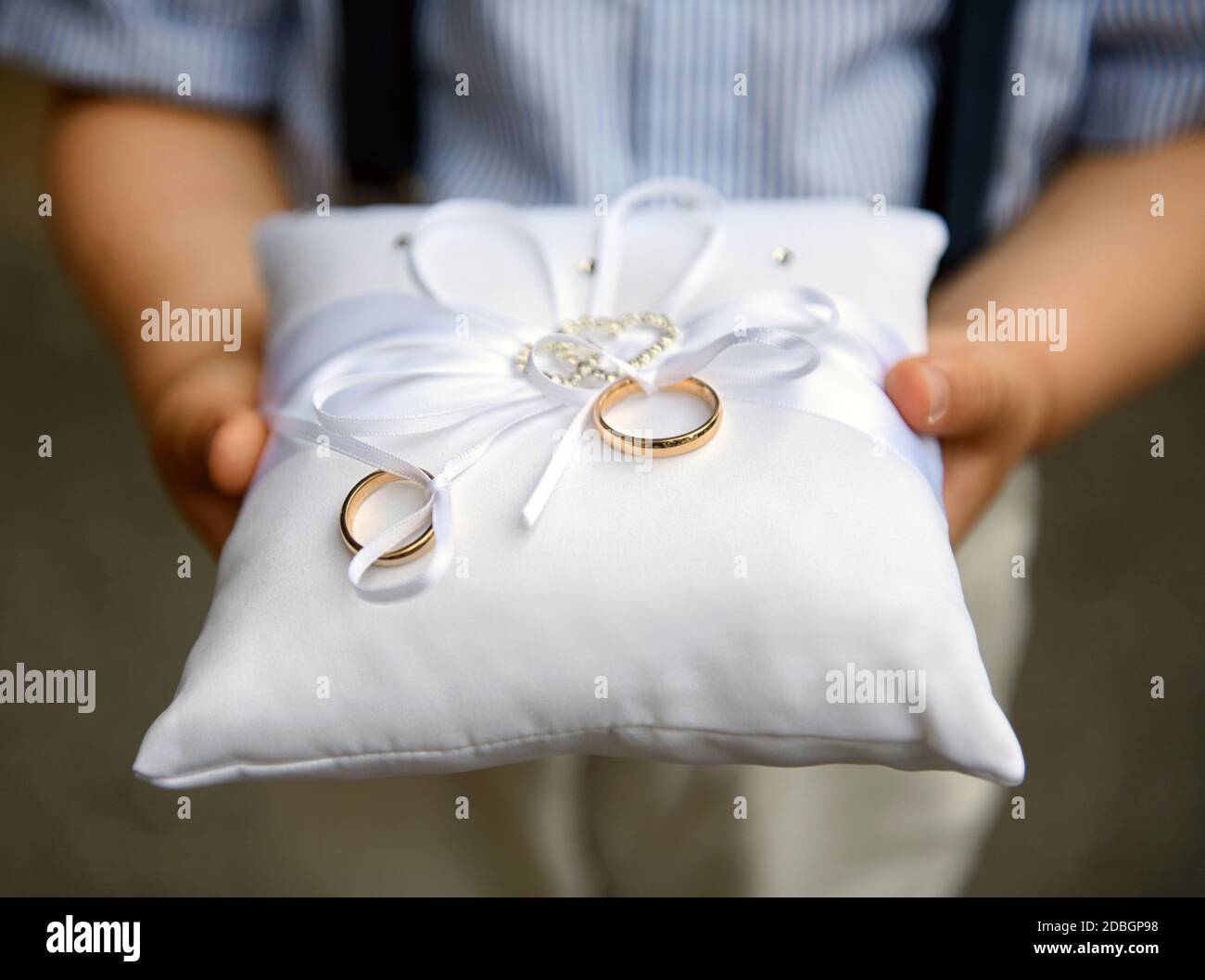 Pageboy che tiene due bande o anelli di nozze d'oro su un cuscino decorato con arco bianco durante la cerimonia di matrimonio a. una chiesa in primo piano Foto Stock