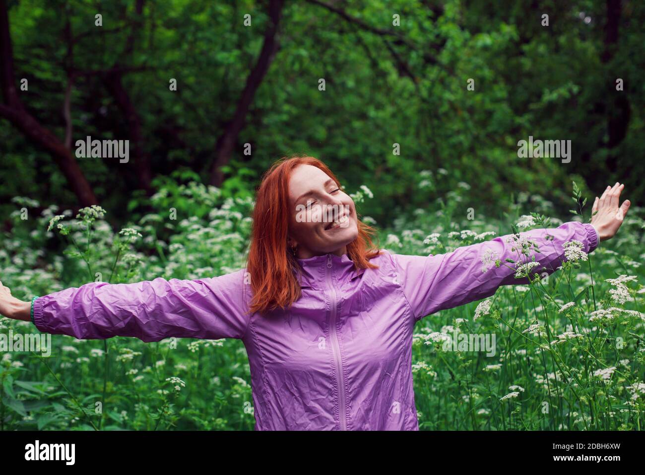 Donna rossa felice con mani sparse in piedi in una foresta con occhi chiusi e sorriso gioioso. Foto Stock