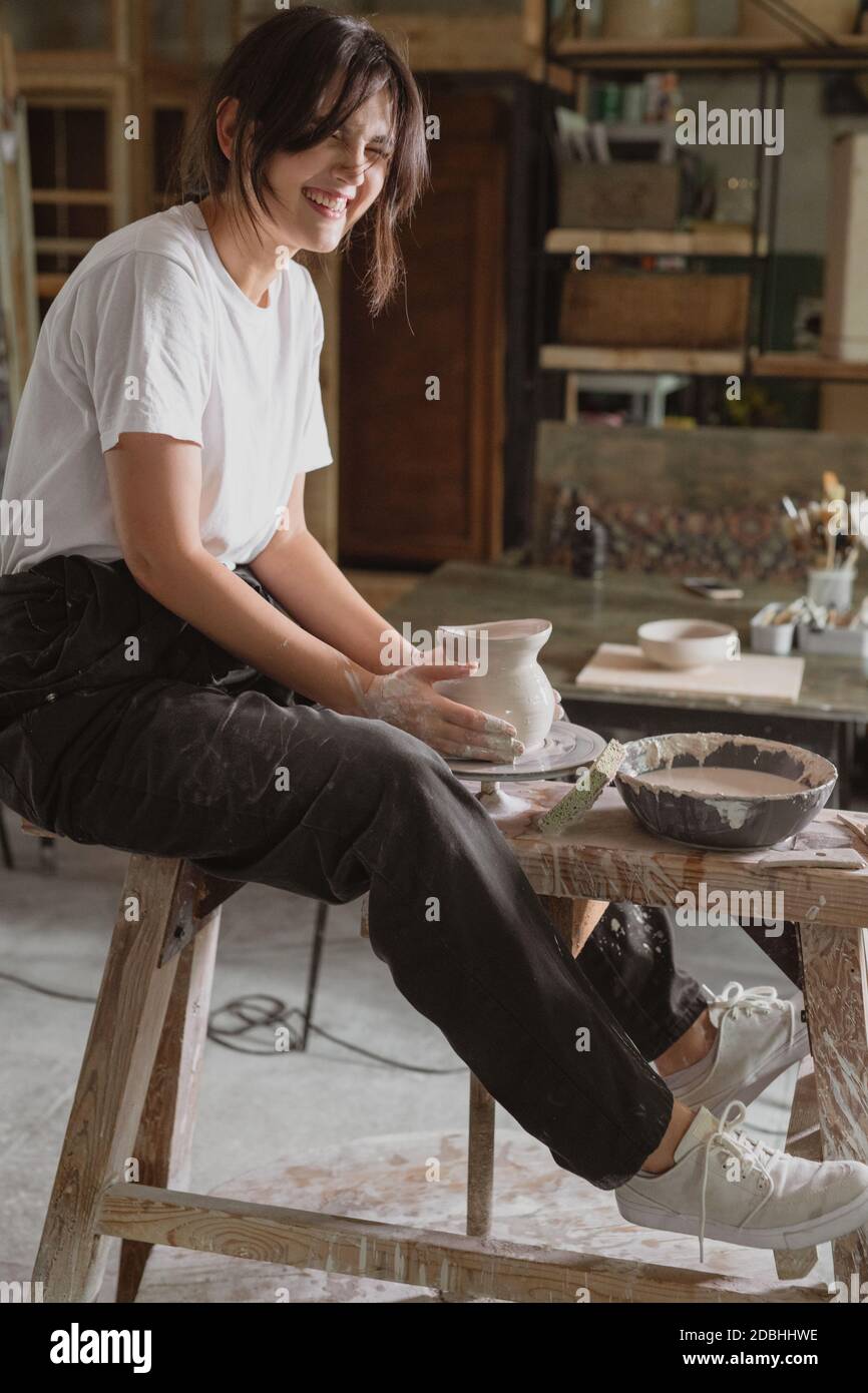 Bella donna ridente che lavora sulla ruota dei vasai facendo brocca di argilla in laboratorio di ceramica. Foto Stock
