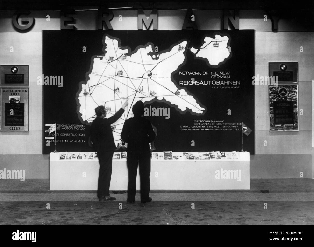 Al London Motor Show nella Sala Olimpica, un poliziotto inglese è mostrato una mappa panoramica della rete tedesca di Reichsautobahnen (autostrade) emessa dal Reichsbahnzentrale fuer den deutschen Reiseverkehr. Le autostrade attualmente in fase di completamento e le autostrade proposte sono tutte indicate sulla mappa. A sinistra e a destra ci sono i consigli di amministrazione di BMW, Auto Union e Mercedes-Benz. Foto Stock