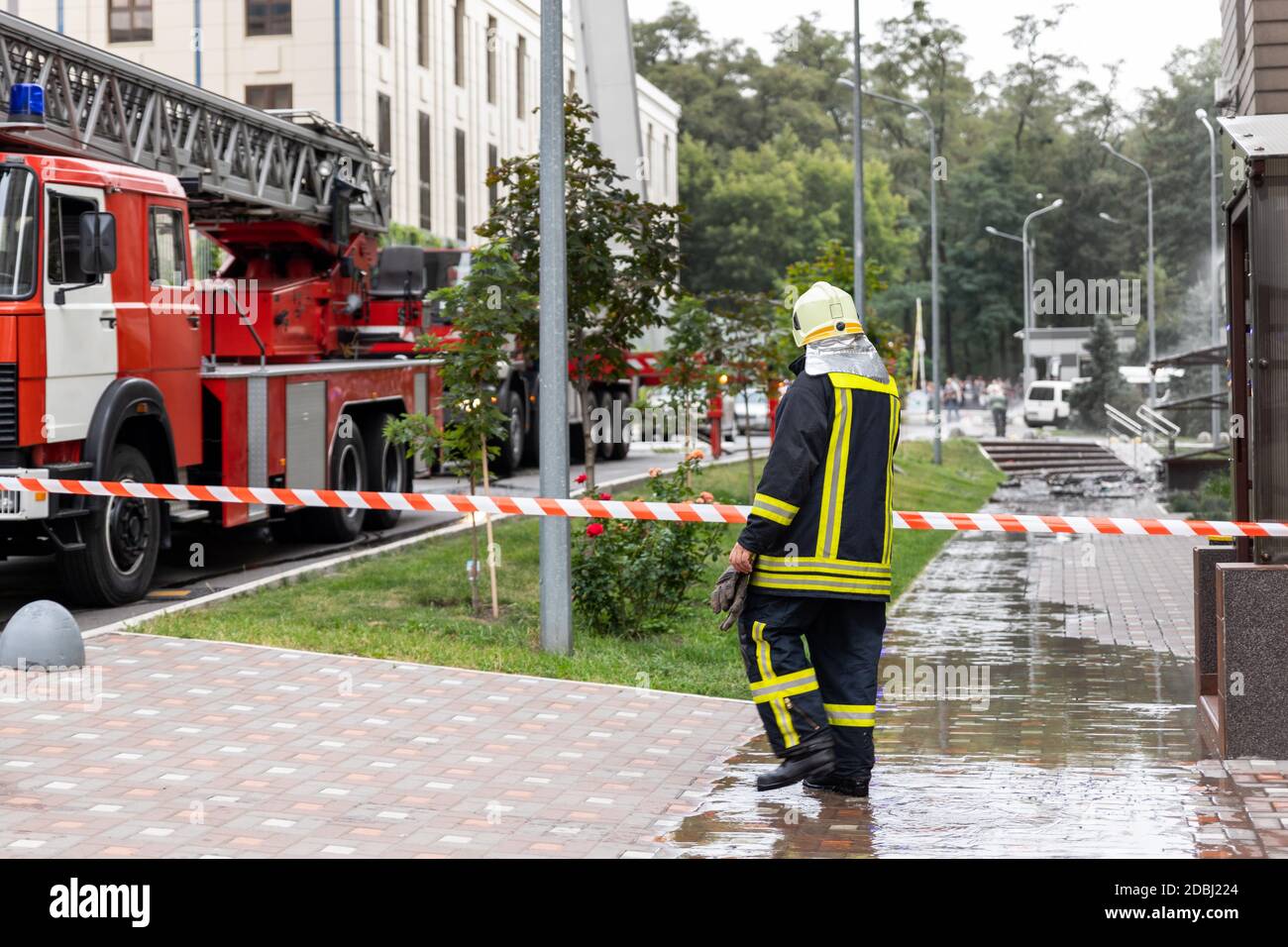 Vigili del fuoco che indossano uniforme in piedi dietro nastro antincendio e molti incendi carrelli motore con scaletta in caso di incidente nella torre alta residenziale o edificio di uffici Foto Stock