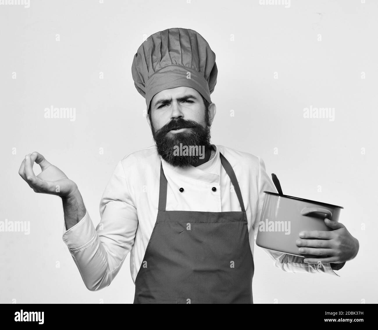 Lo chef tiene una zuppa o una composta facendo gesti italiani. Cuoco con simpatico grimace in uniforme borgogna ha casseruola con mestolo. L'uomo con la barba tiene una pentola rossa su sfondo bianco. Concetto di cucina professionale Foto Stock