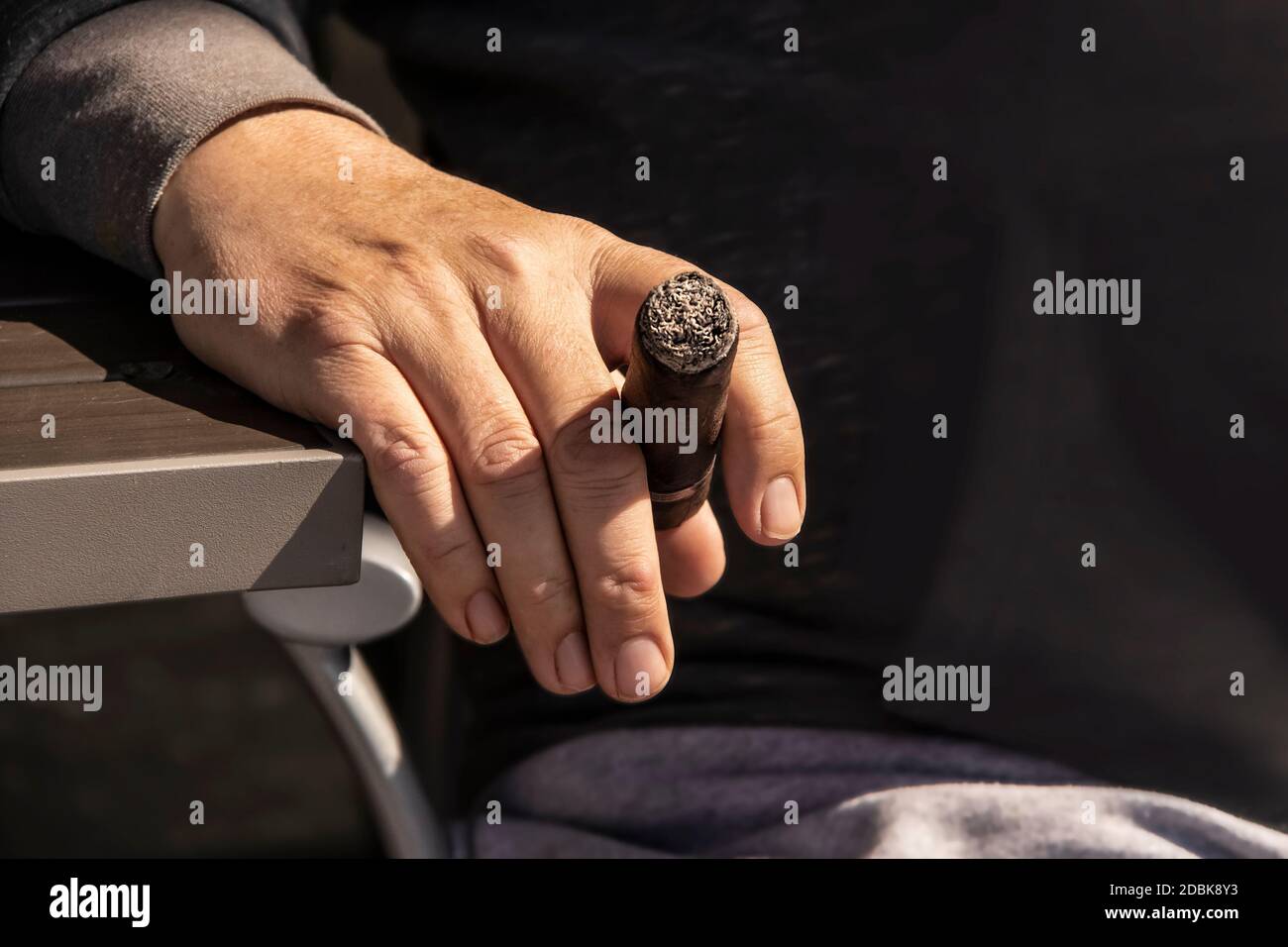 Primo piano della mano dell'uomo che poggia sul lato del tavolo accanto la sua sedia che tiene un sigaro illuminato Foto Stock