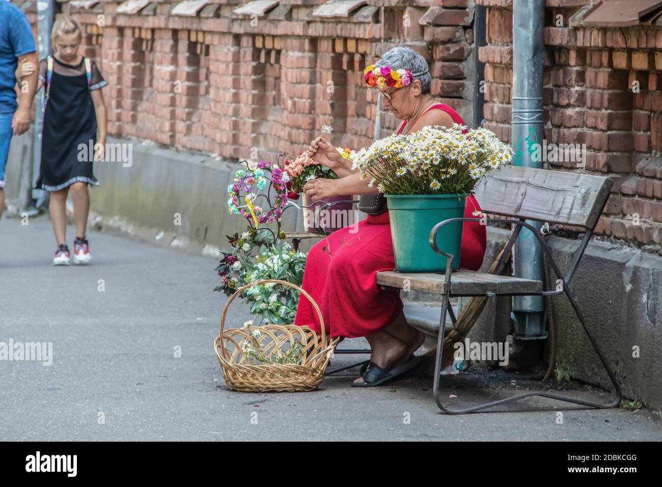 7-15 2019 Tbilisi Georgia - una donna anziana in A. dress siede su una panchina da un muro di mattoni e. fa le testate dai fiori freschi da vendere al turistico Foto Stock