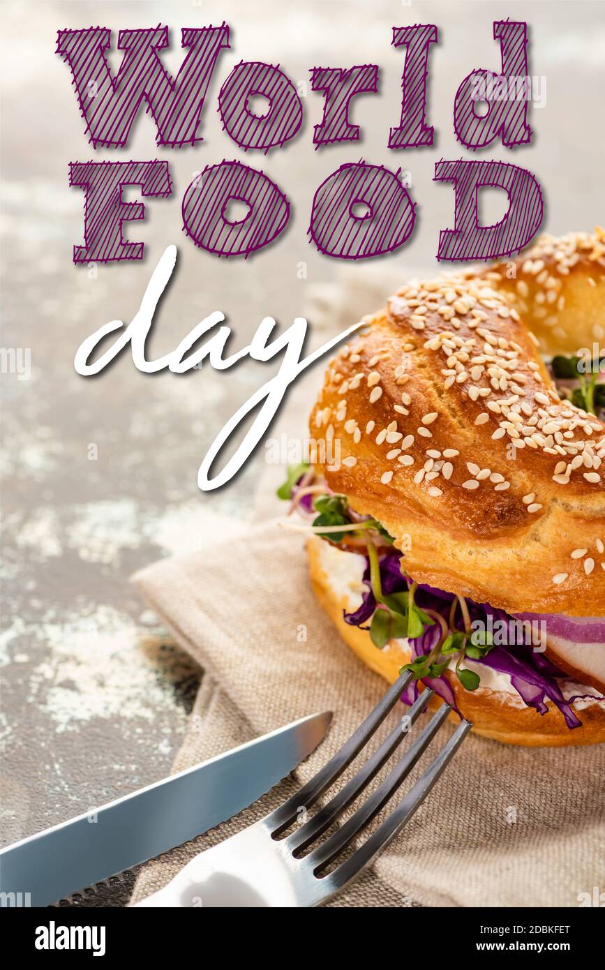 saporito bagel con carne su tovagliolo con posate vicino al mondo scritta food day su superficie testurizzata Foto Stock