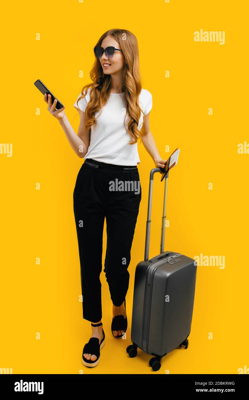 Una giovane donna felice in occhiali scuri, che porta una valigia e un passaporto con i biglietti, utilizza un telefono cellulare su sfondo giallo. Il concetto di viaggio, Foto Stock