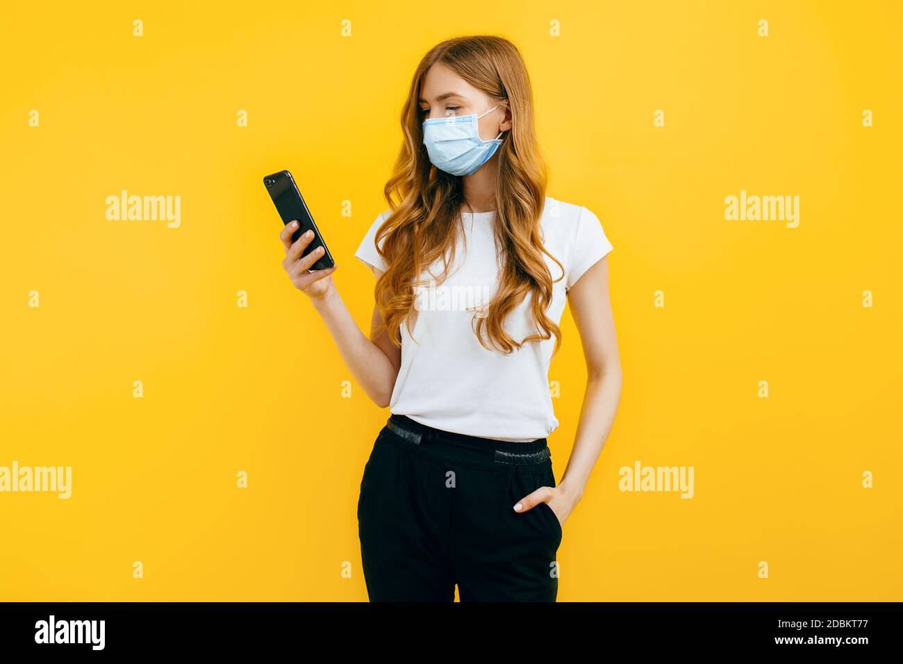 Una giovane donna, indossando una maschera medica protettiva, utilizza un telefono cellulare su uno sfondo giallo. Il concetto di quarantena, coronavirus Foto Stock