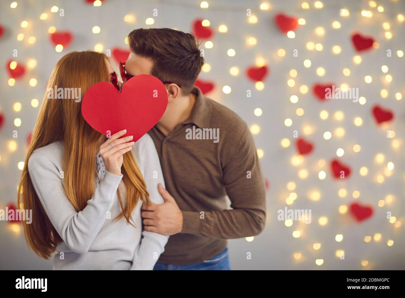 Giovane coppia felice in amore in piedi e baciando dietro grande cuore rosso nelle sue mani Foto Stock