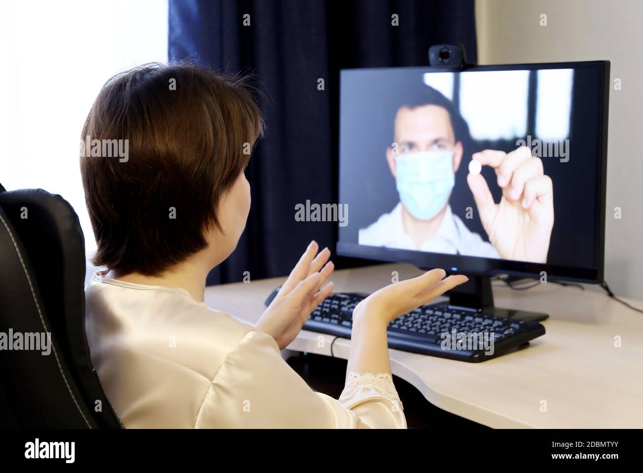 Consultazione online con un medico, donna ascolta le raccomandazioni del medico seduto di fronte alla webcam del PC. Uomo in maschera medica con pillola Foto Stock