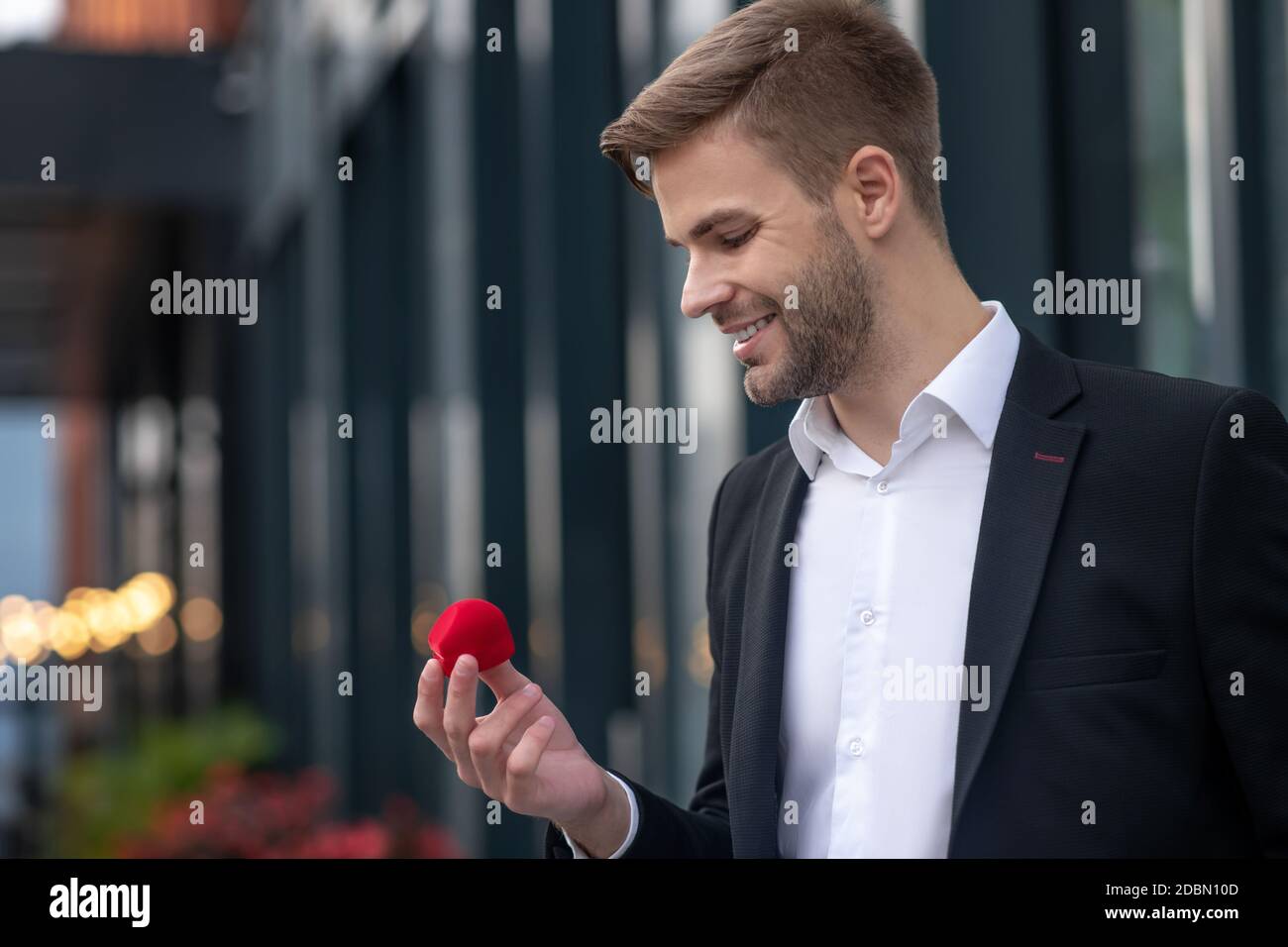 Giovane bell'uomo in tuta nera che tiene una scatola con anello di fidanzamento e aspetto eccitato Foto Stock