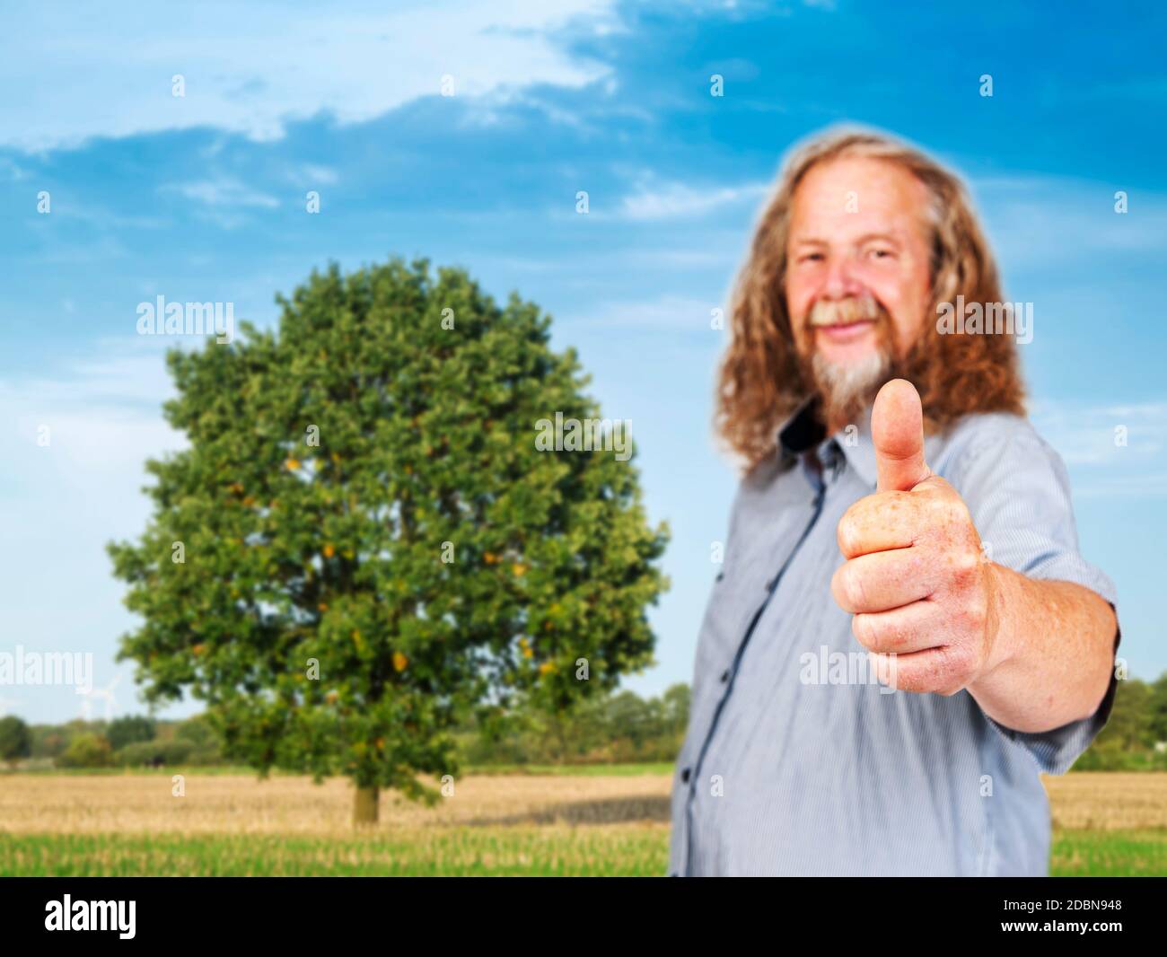 Vista laterale del corpo superiore di un uomo dai capelli lunghi, di fronte ad un unico albero deciduo con una grande cima d'albero su terreni agricoli estivi nel blurr Foto Stock