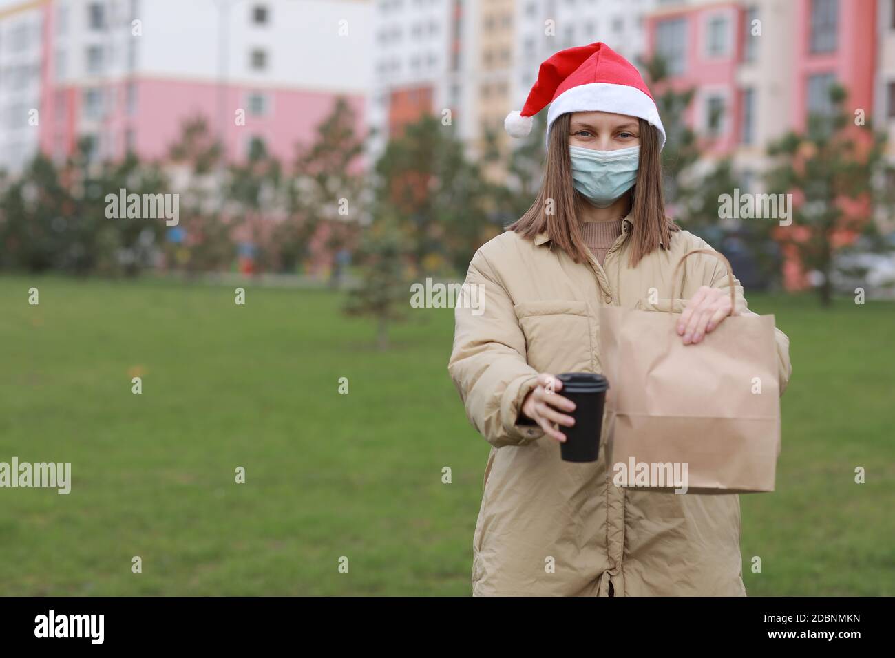 Ragazza Courier in maschera protettiva viso e cappello di babbo natale  tiene una borsa di carta con prodotti e una tazza di caffè all'aperto.  quarantena di natale coronavirus, s Foto stock -
