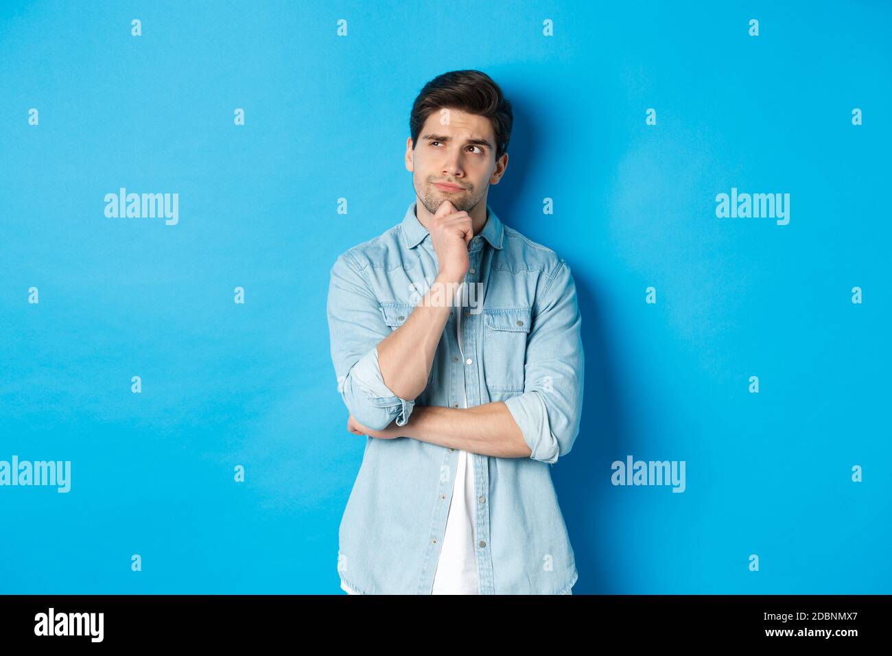 Immagine di un uomo adulto sopportato 25 anni, pensando a qualcosa, guardando l'angolo in alto a sinistra e meditando idee, in piedi su sfondo blu Foto Stock