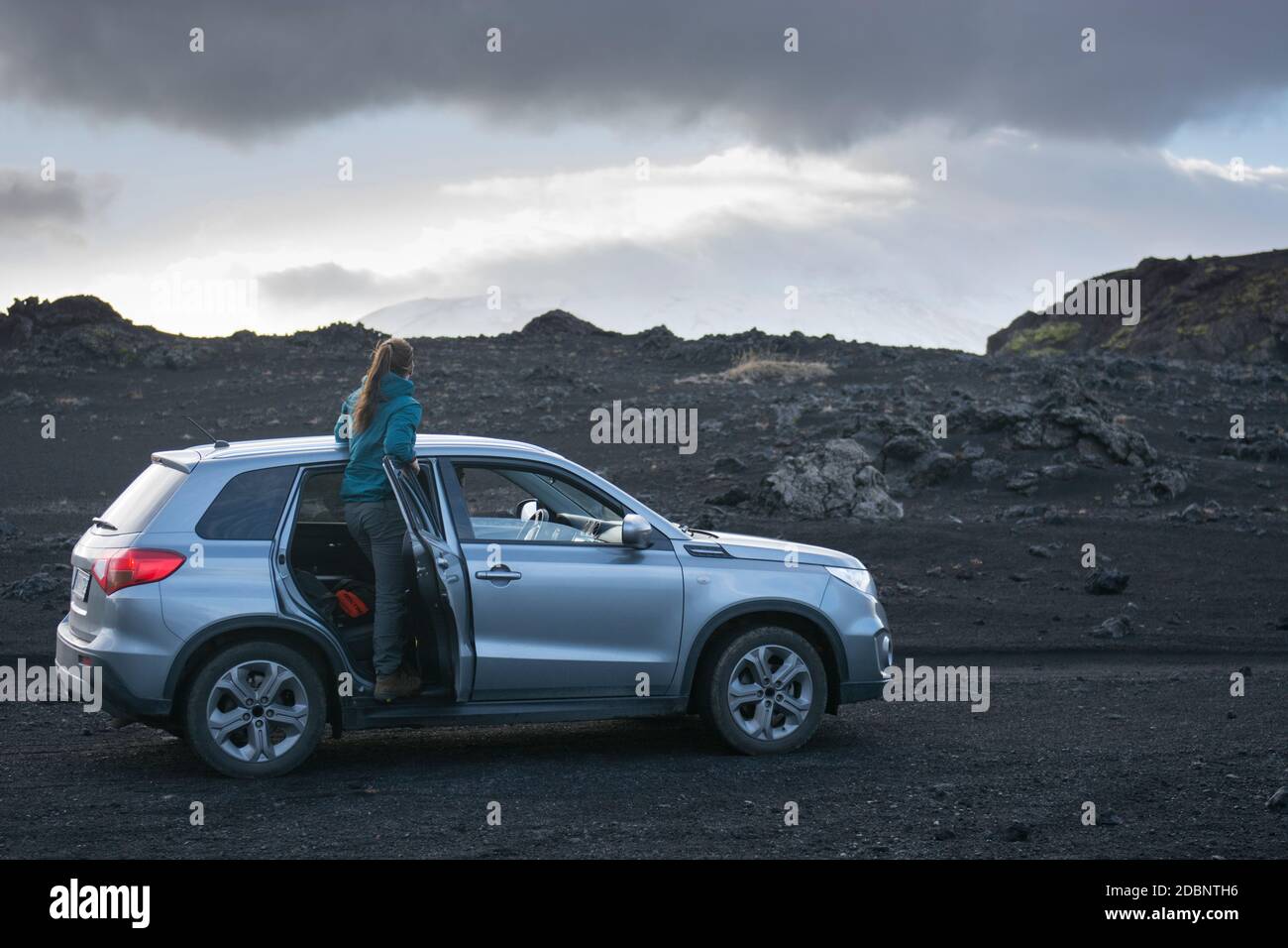 Una giovane donna avventurosa con una giacca blu, si appoggia da un SUV d'argento mentre si allontana in lontananza, guardando la montagna innevata coperta da c Foto Stock