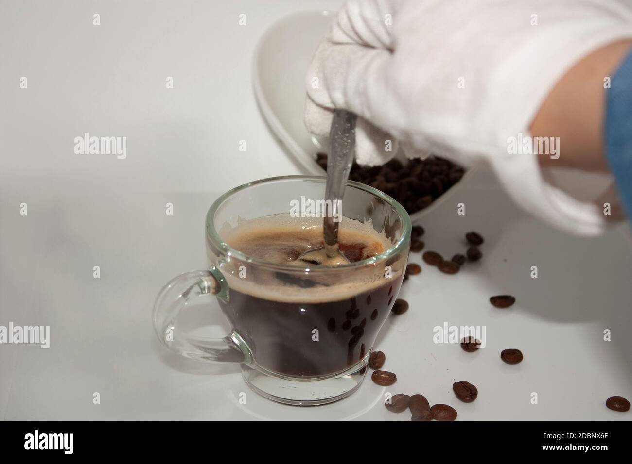 Tazzina per la cottura a vapore con caffè e schiuma che si muove agitando il cucchiaio con l'aiuto di una mano bianca con guanti, accanto a un bolo bianco a forma di cuore in ceramica Foto Stock