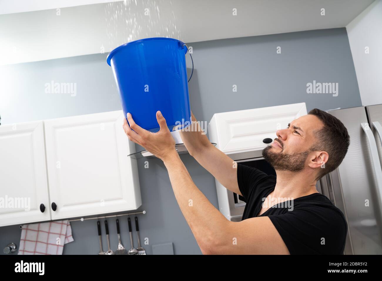 Benna di tenuta dell'uomo preoccupata mentre goccioline di acqua fuoriescono dal soffitto in cucina Foto Stock