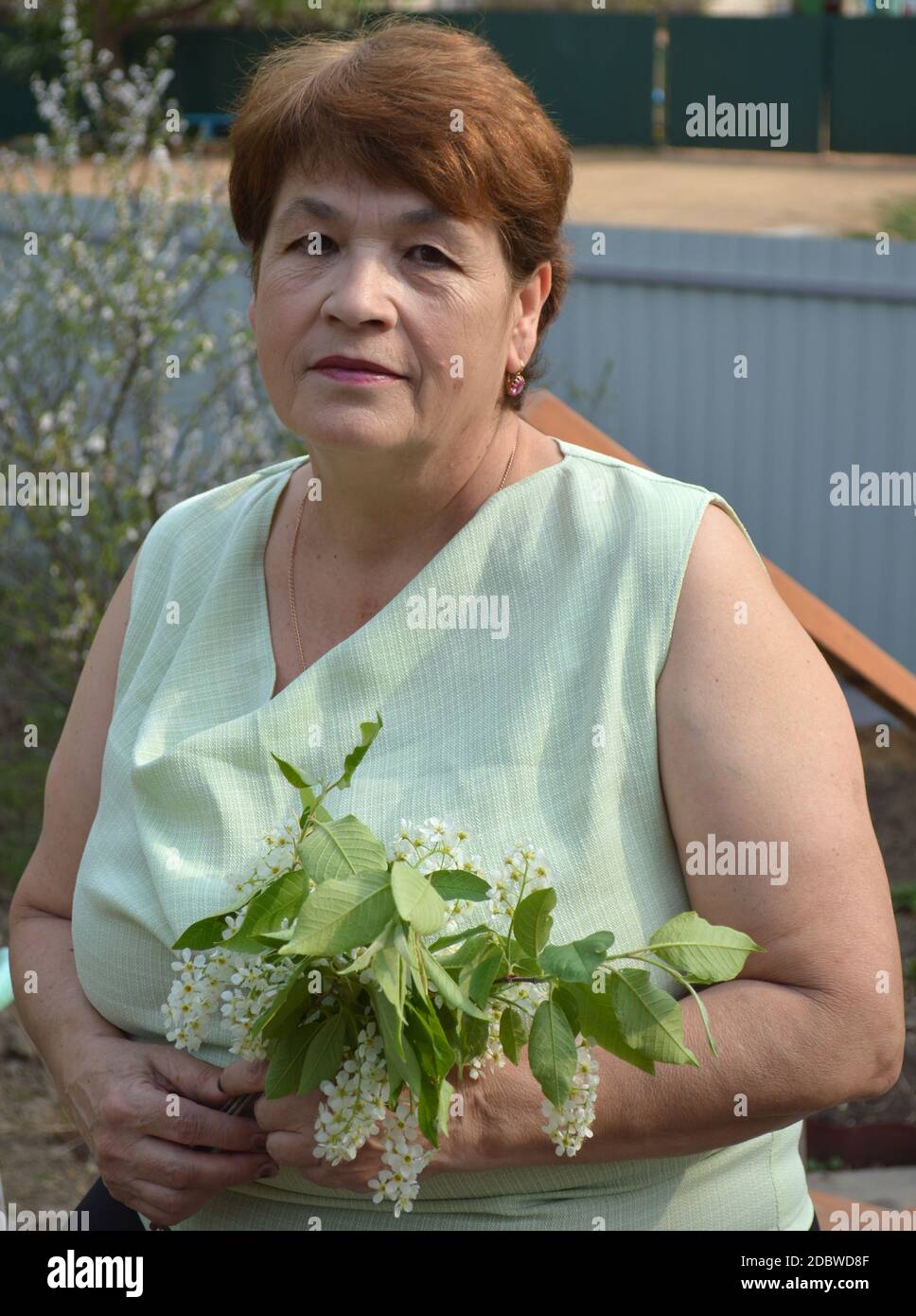 Ritratto di 65 y.o. donna anziana in blusa senza maniche con espressione facciale neutra seduta e tenendo il bouquet primaverile di rami fioriti Foto Stock