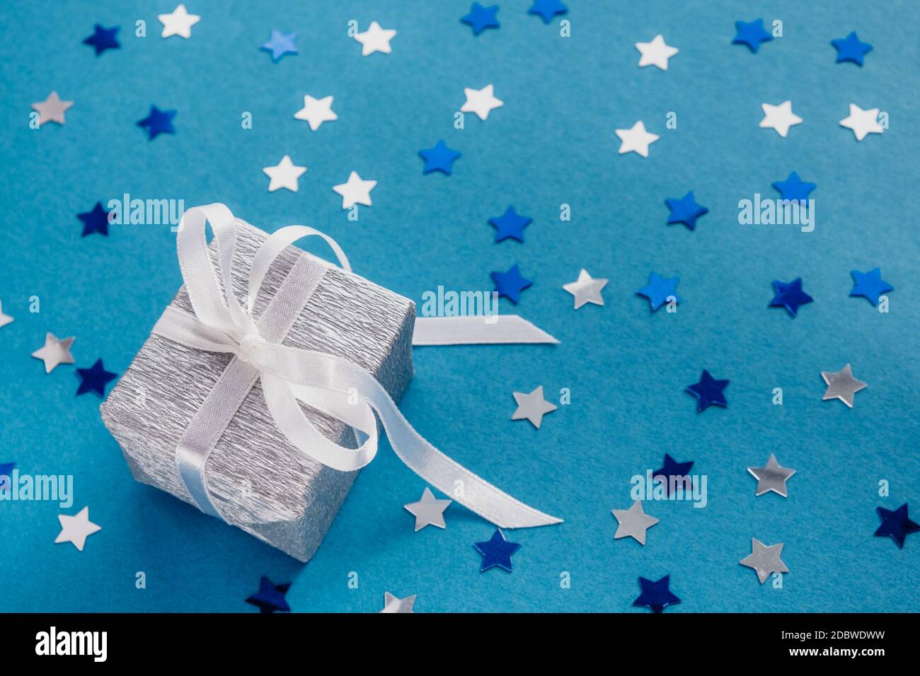 Celebration card con scatola regalo argentata con nastro bianco, su sfondo blu con stella confetti. Messa a fuoco selettiva. Primo piano. Spazio di copia. Foto Stock