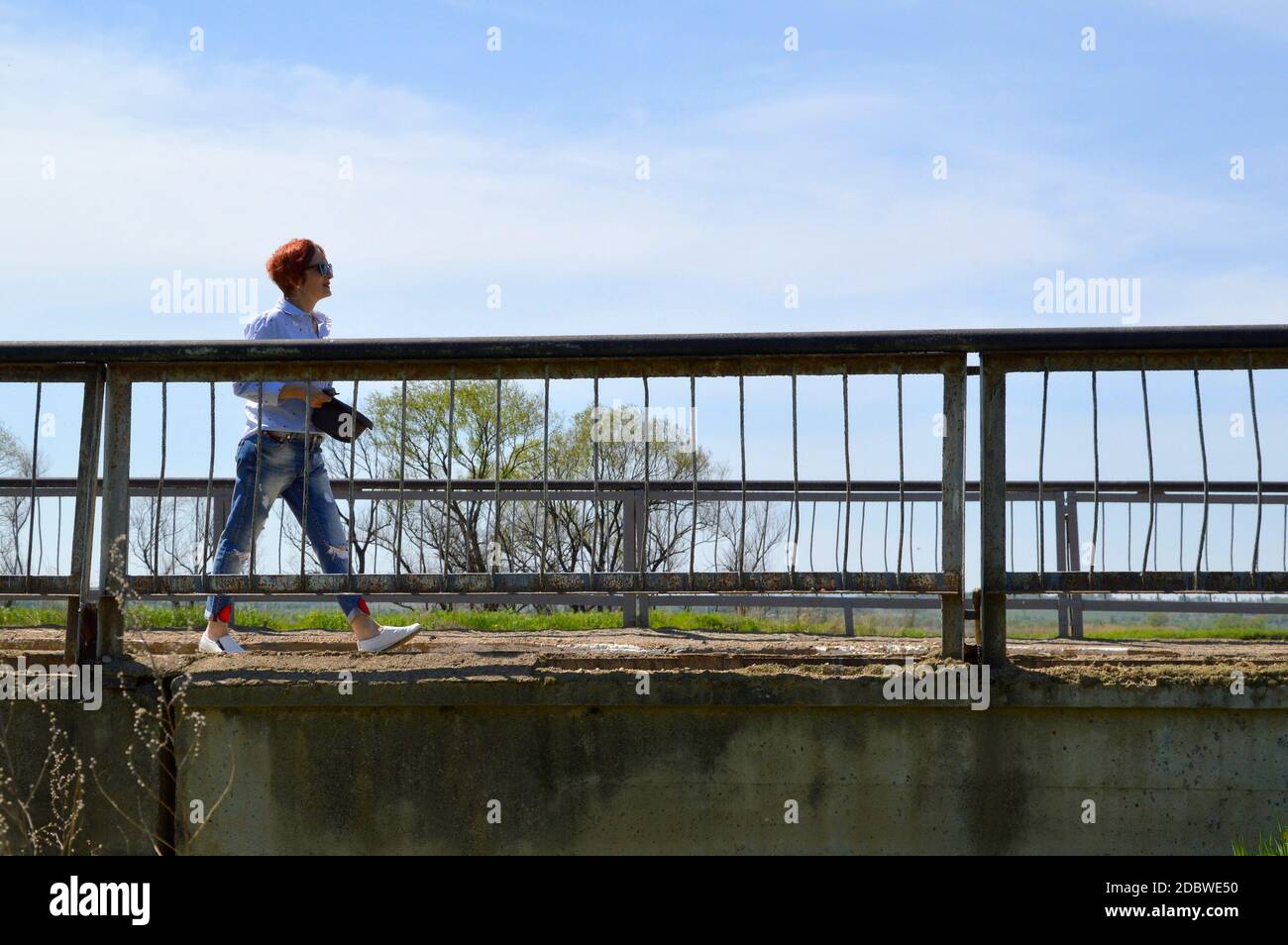 Donna dai capelli rossi con un cappello che passa sopra il ponte e che guarda via in una giornata estiva. Spazio copia Foto Stock