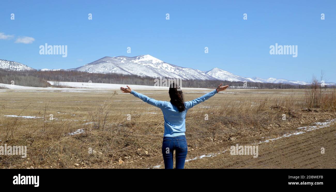 Donna di bruna snella che abbraccia la natura a braccia aperte. Il piede delle montagne innevate. Concetto di uscire dopo la quarantena. Russia Foto Stock