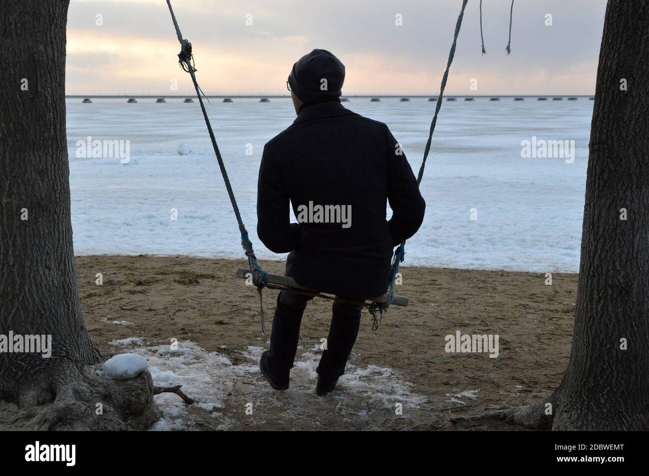 L'uomo adulto in un cappotto oscilla su un'oscillazione e. guarda attentamente il mare in una fredda serata Foto Stock