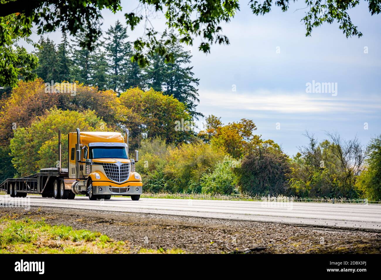 Carro lungo giallo brillante Big RIG rosso industriale semi-camion trasporto di un semirimorchio a gradino vuoto in marcia rettilinea strada autostradale con verde Foto Stock