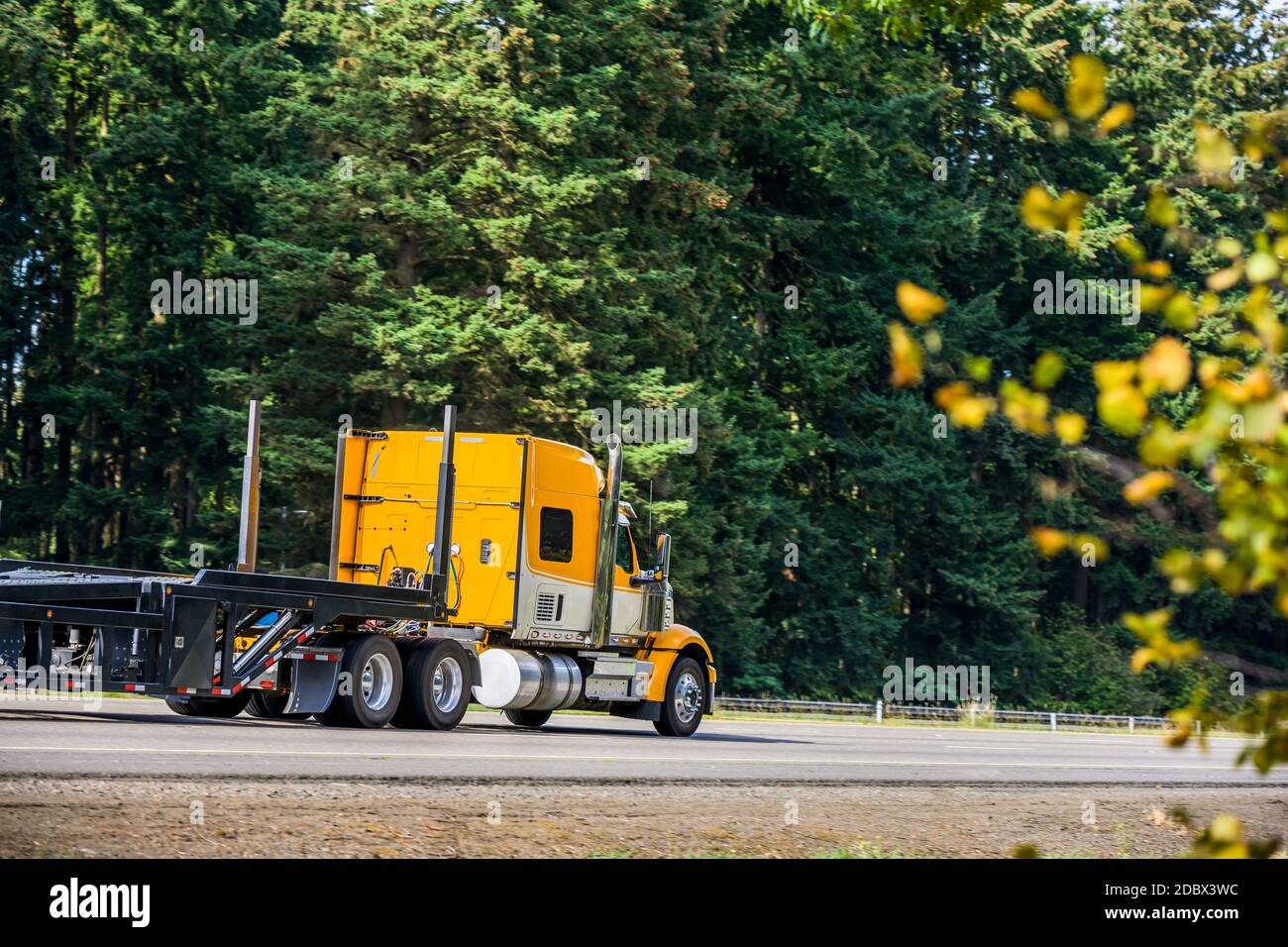 Carro lungo giallo brillante Big RIG rosso industriale semi-camion trasporto di un semirimorchio a gradino vuoto in marcia rettilinea strada autostradale con verde Foto Stock