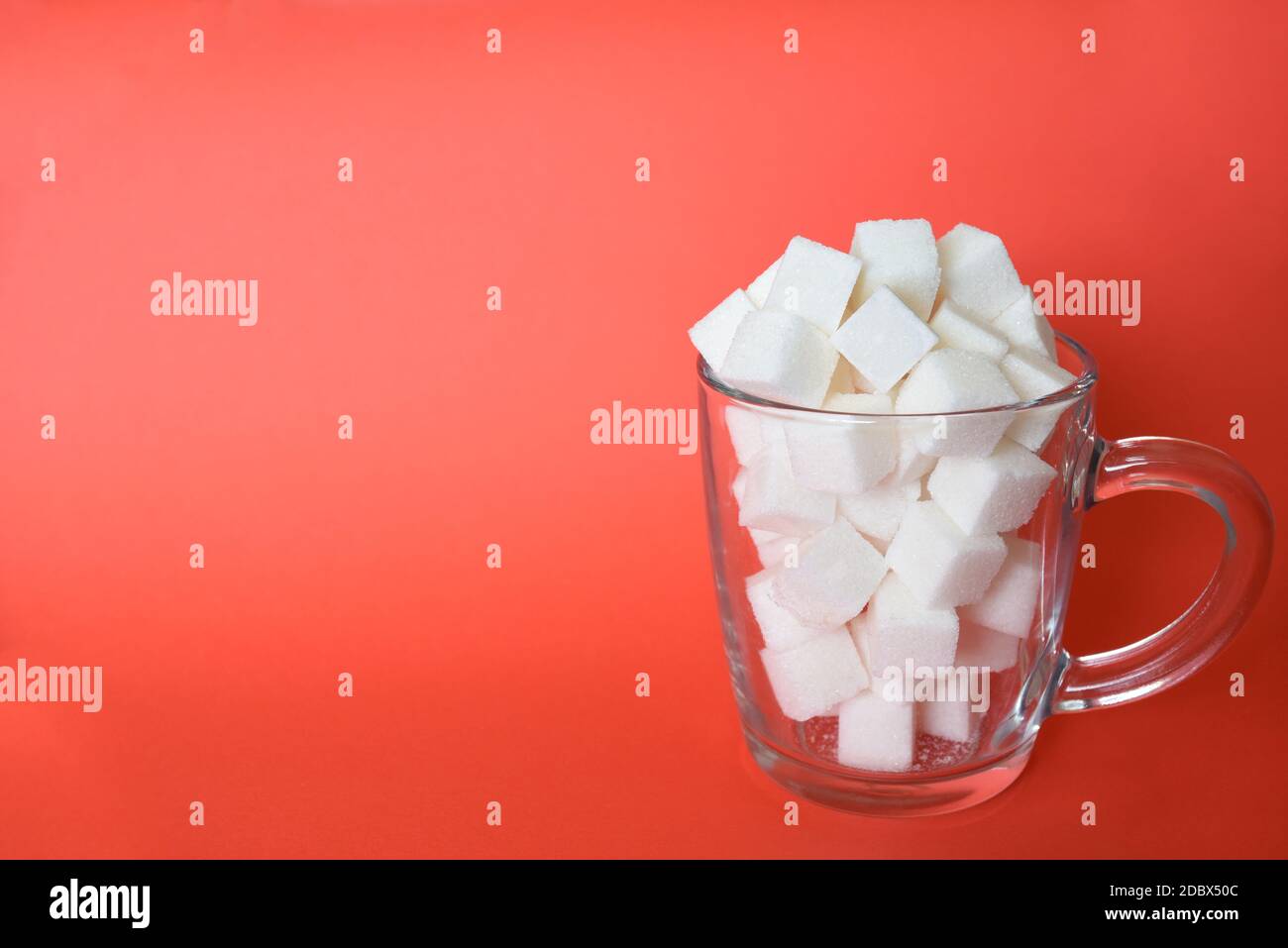 Tazza trasparente piena di raffinati cubetti di zucchero bianco su sfondo rosso. Spazio di copia Foto Stock