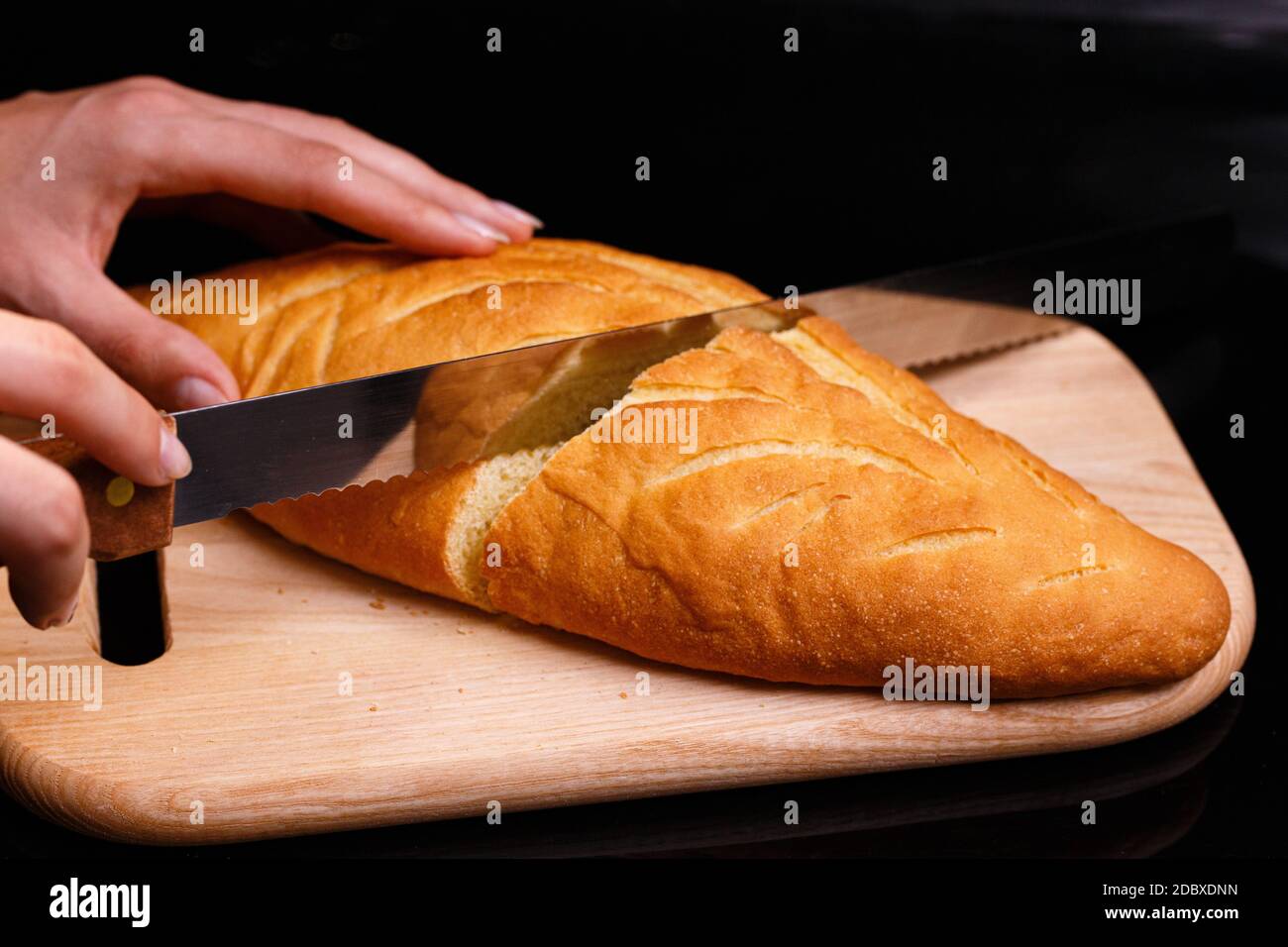 Il pane bianco fatto in casa viene tagliato con un grande coltello. Cottura a casa. Pane allungato su una tavola di legno. Foto su sfondo nero. Foto Stock