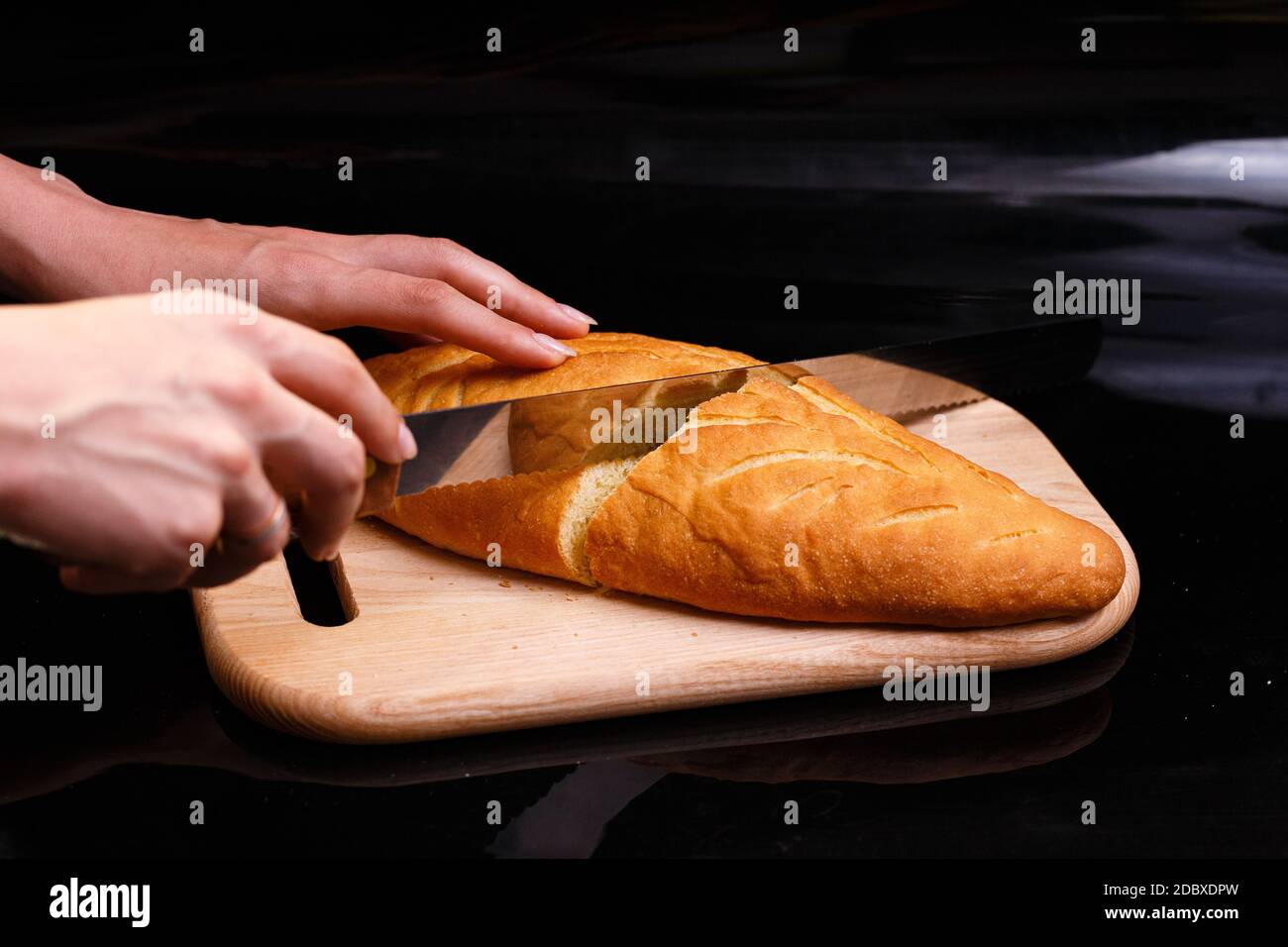 Il pane bianco fatto in casa viene tagliato con un grande coltello. Cottura a casa. Pane allungato su una tavola di legno. Foto su sfondo nero. Foto Stock