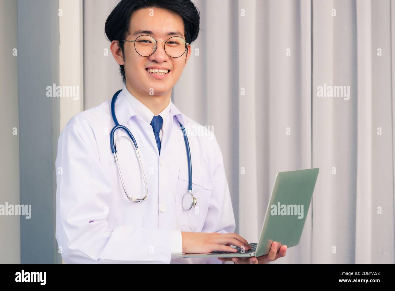 Ritratto del giovane bel dottore asiatico che indossa un vestito medico e stetoscopio sorridente seduto in ospedale, concetto di assistenza sanitaria Foto Stock
