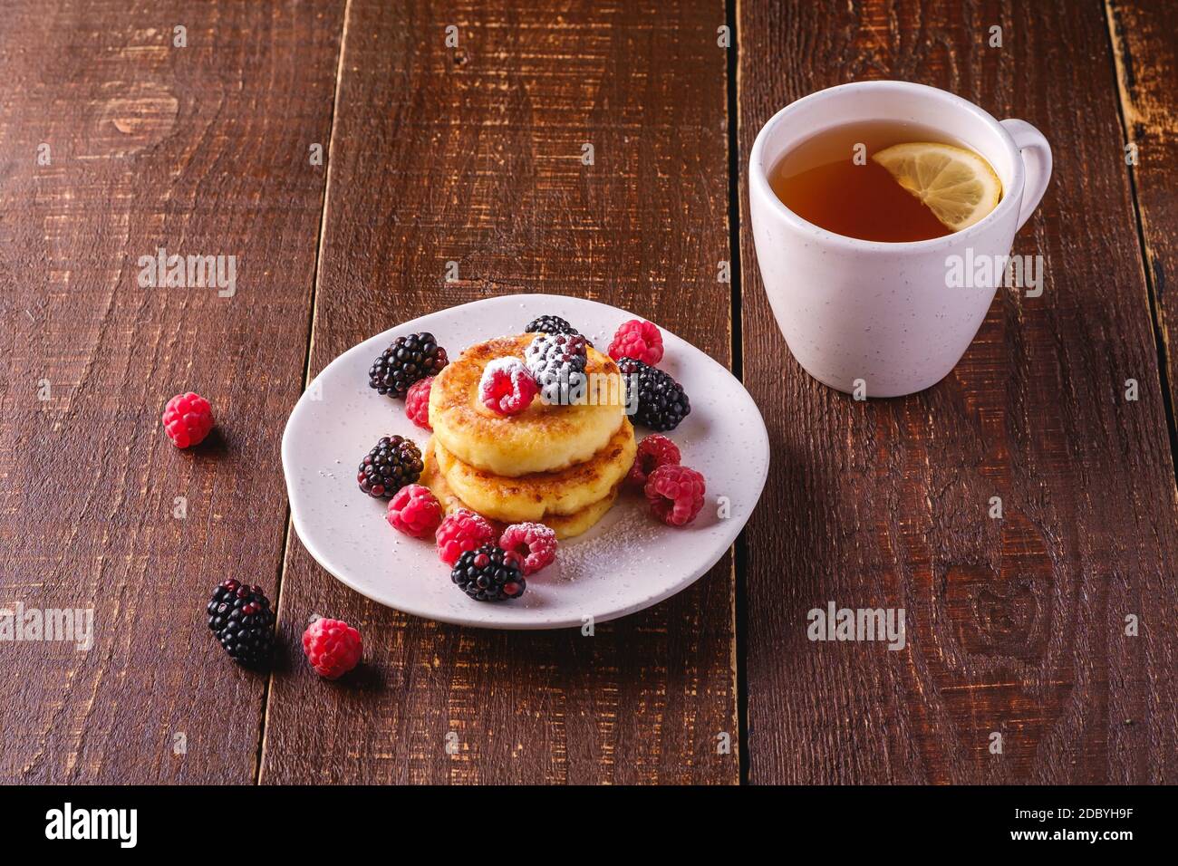 Frittelle di formaggio cottage, frittelle di cagliata dessert con lampone e frutti di bosco in piatto vicino alla tazza di tè caldo con fetta di limone Foto Stock
