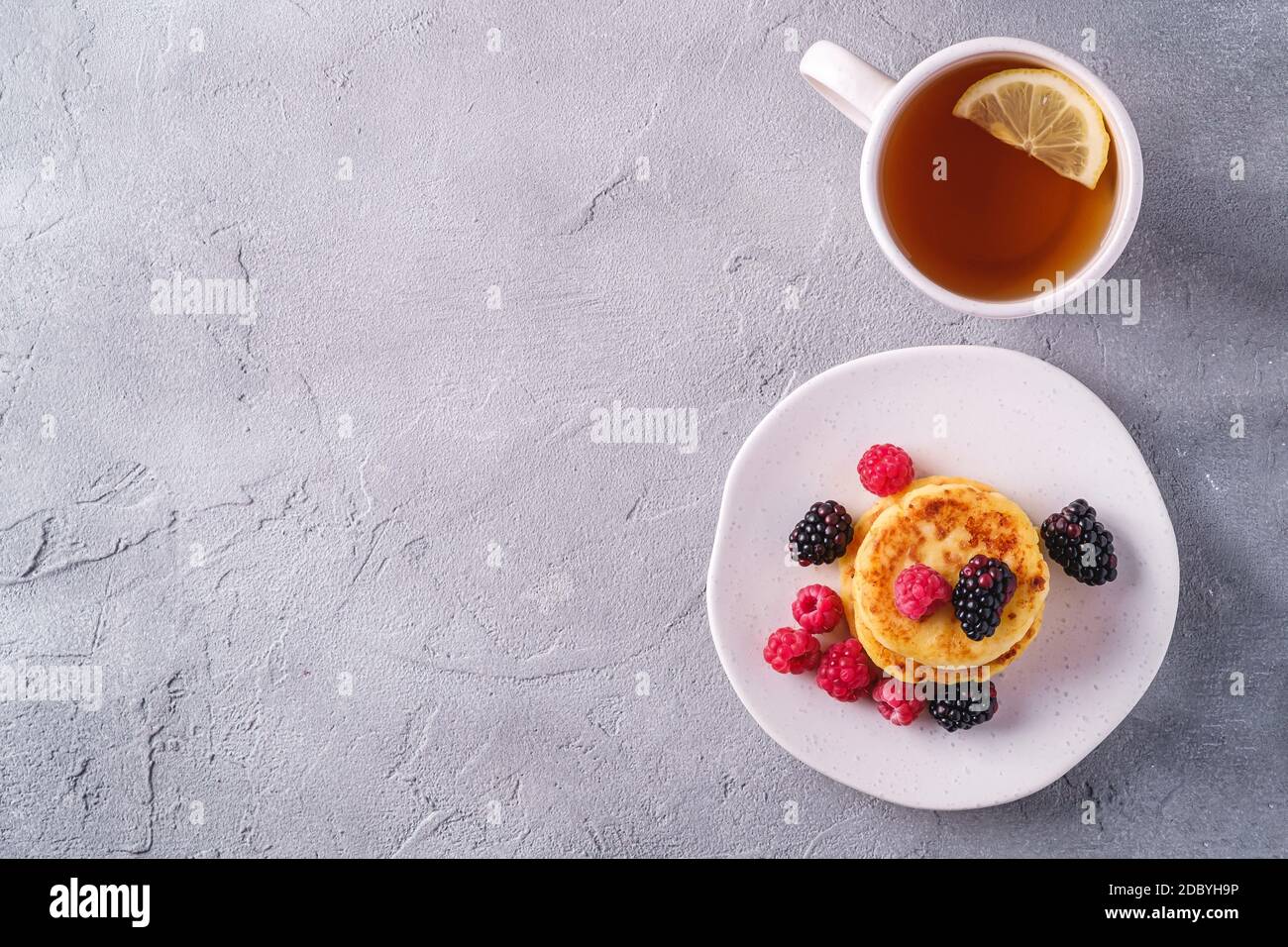 Frittelle di formaggio cottage, frittelle di cagliata dessert con lampone e bacche di mora in piatto vicino alla tazza di tè caldo con fetta di limone su cemento di pietra Foto Stock