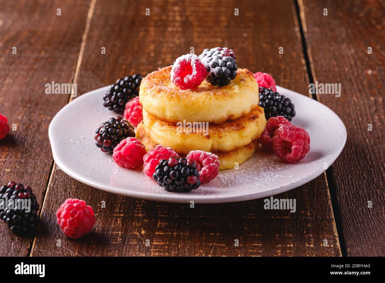 Frittelle di formaggio e zucchero in polvere, frittelle di cagliata dessert con lampone e frutti di bosco in piatto su sfondo di legno marrone scuro Foto Stock