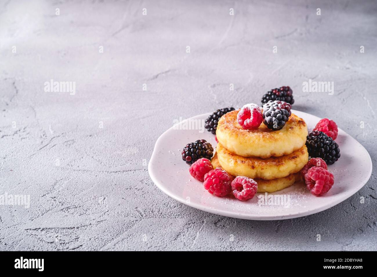 Frittelle al formaggio di cottage e zucchero in polvere, frittelle di cagliata dessert con lampone e bacche di mora in piastra su fondo di cemento di pietra Foto Stock