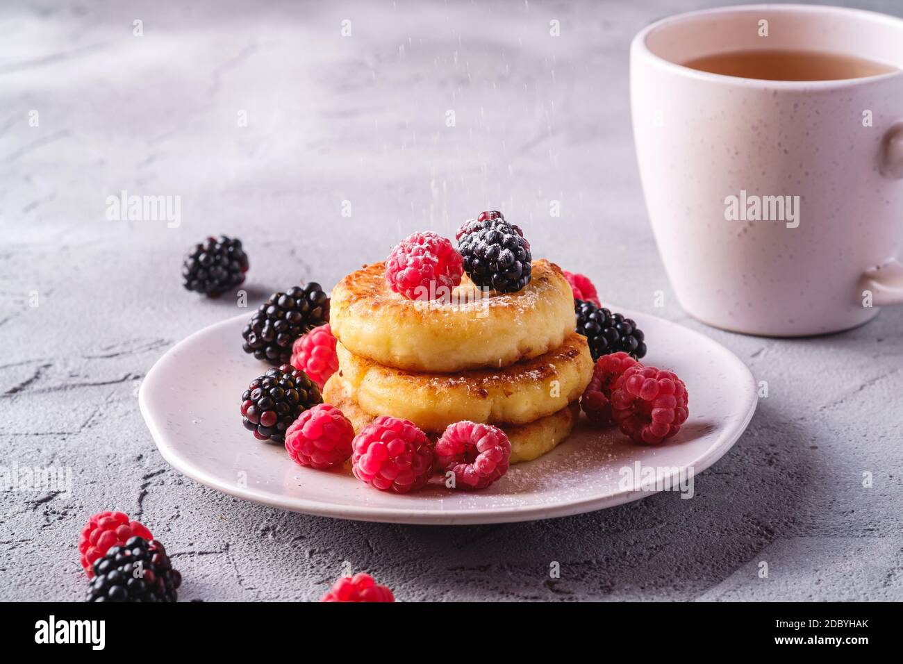 Frittelle al formaggio e zucchero in polvere, frittelle di cagliata dessert con lampone e frutti di bosco in piatto vicino alla tazza di tè caldo con fetta di limone Foto Stock
